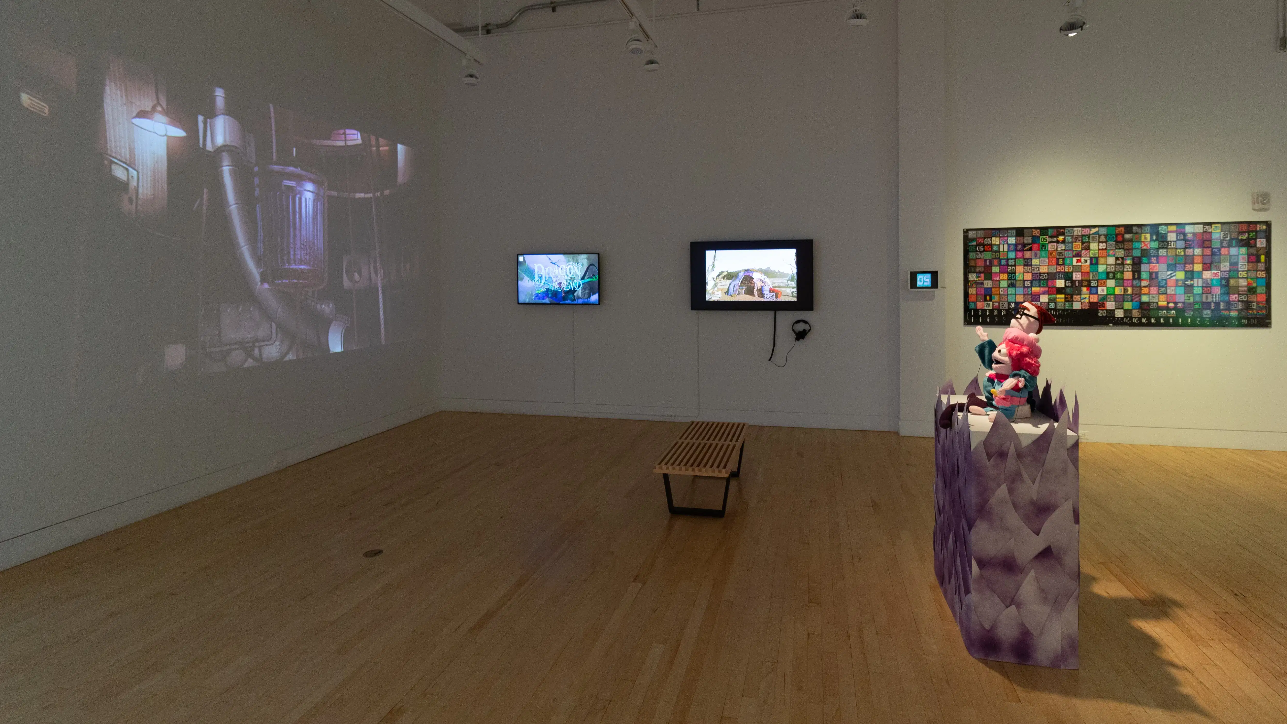 Installation image featuring a projection to the left, two monitors and a print on the back wall, and a pedestals with plush sculptures in the foreground.