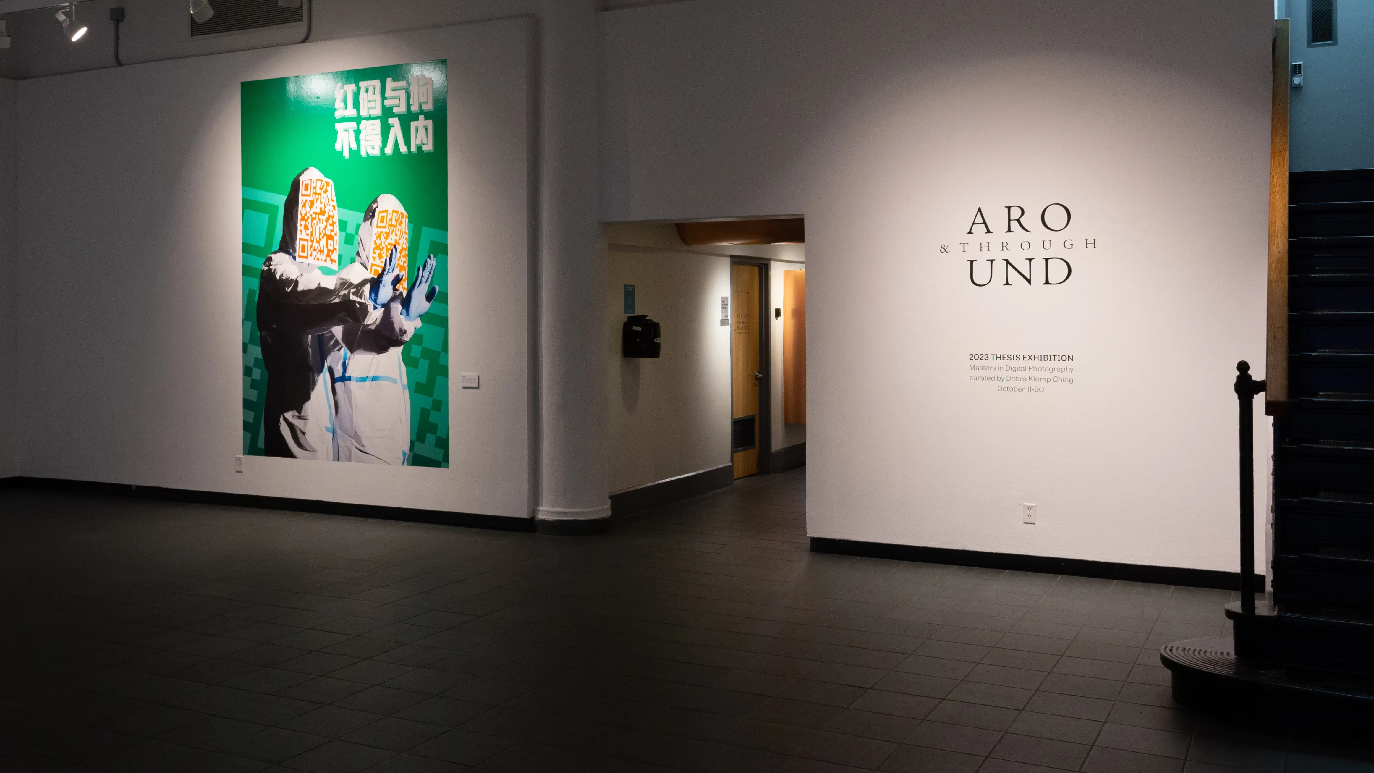 A hallway cuts through one wall of the gallery, with a large bright green image showing two people in quarantine gear and QR code-covered faces to the left of the doorway. On the right, wall vinyl reads “Around & Through 2023 Thesis Exhibition”.