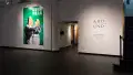 A hallway cuts through one wall of the gallery, with a large bright green image showing two people in quarantine gear and QR code-covered faces to the left of the doorway. On the right, wall vinyl reads “Around & Through 2023 Thesis Exhibition”.