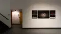 Through a doorway, wall vinyl displays the title of the exhibition. To the right of the doorway, four smaller framed photographs depicting objects against black backgrounds flank either side of one large photograph, all depicting two people against a black background.