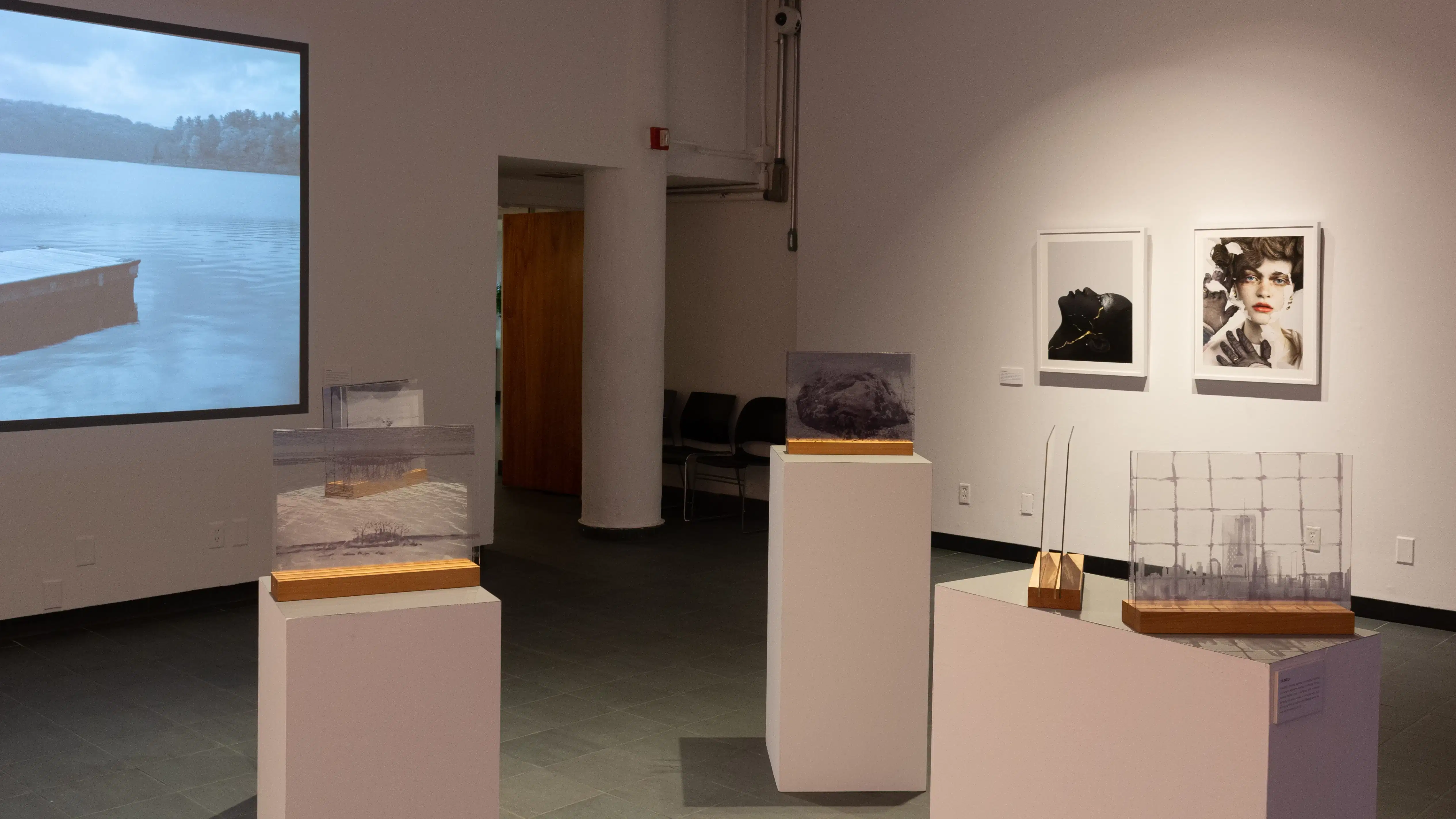 Surrounded by glass pane photographs on pedestals in the center of the gallery, a projection of a video is visible on the wall to the left, and two photographs of people’s faces are visible on the wall to the right.