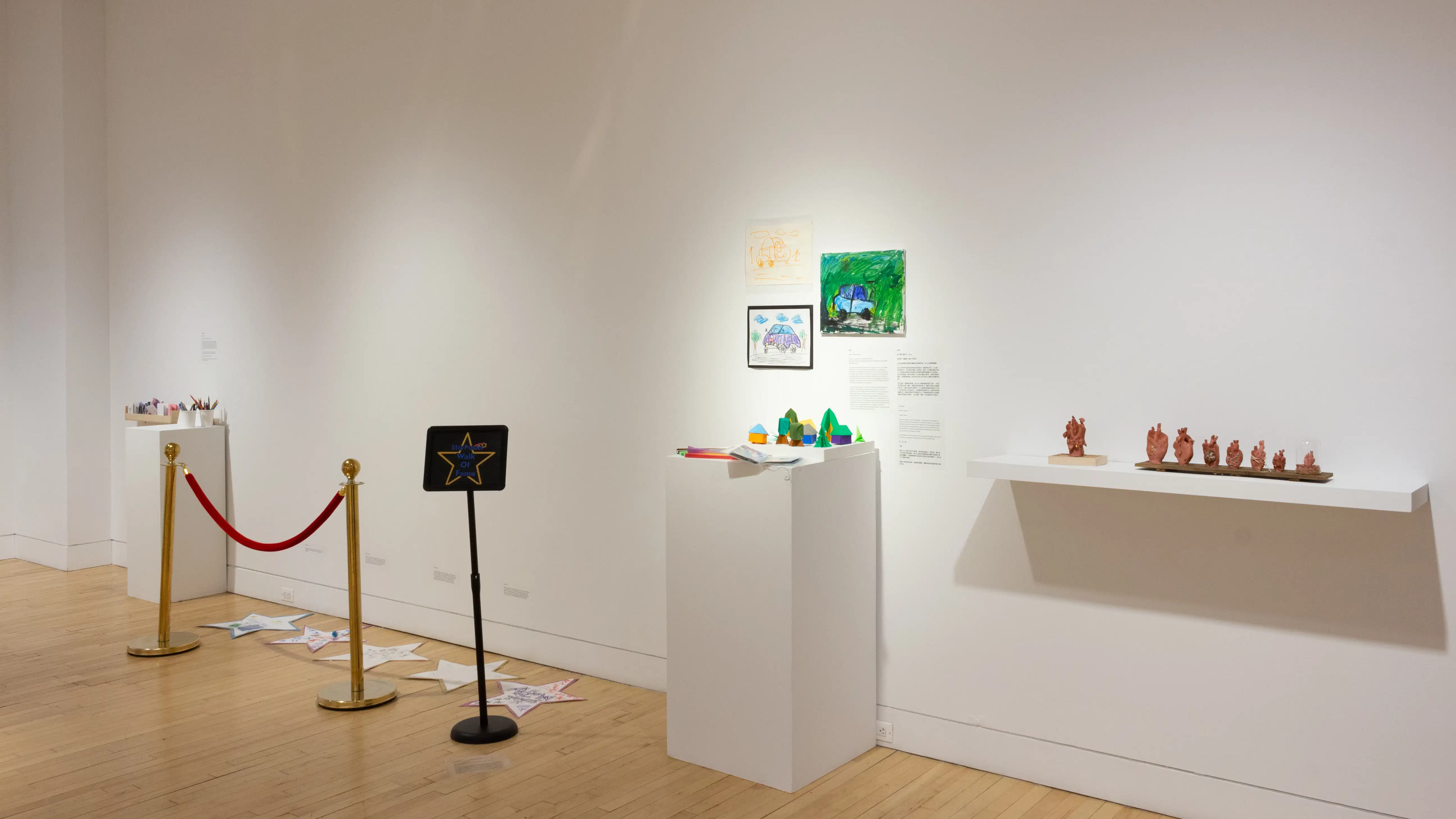 Against one wall of the SVA Flatiron Gallery, a shelf holds several small sculptures of an anatomical heart. To the left of the shelf, colorful paper works sit on and above a pedestal. Further left, a stanchion with a red velvet rope protects a row of paper stars that lay on the ground.