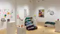 A wider view of the gallery shows a chair with a knit blanket across it, surrounded by pedestals that hold small sculptures. One wall holds colorful drawings and a mirror with a vibrant frame.