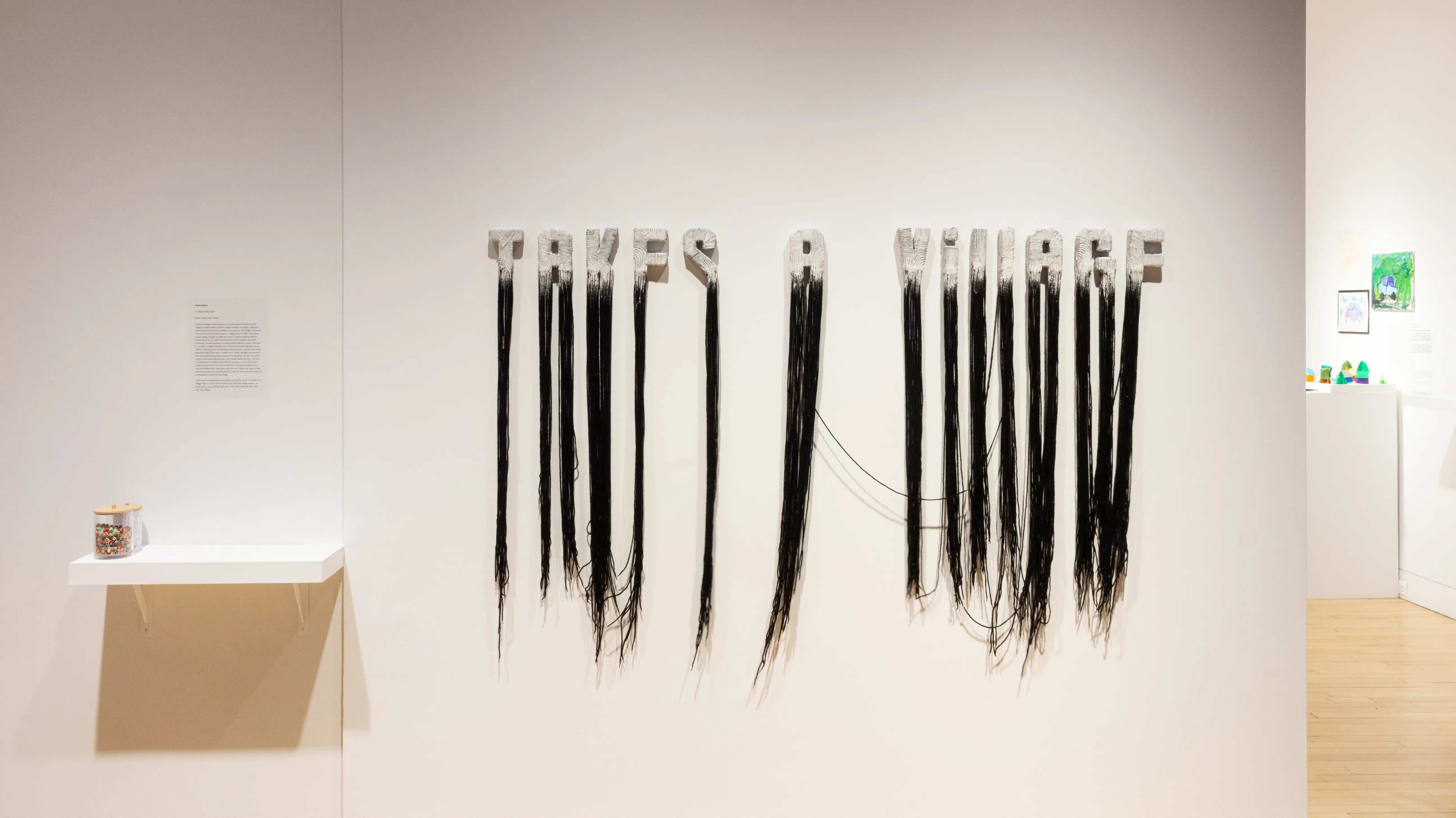 On a wall, the words “TAKES A VILLAGE” are formed out of long black strands, painted white on top. To their left is a jar of beads sitting on a shelf.