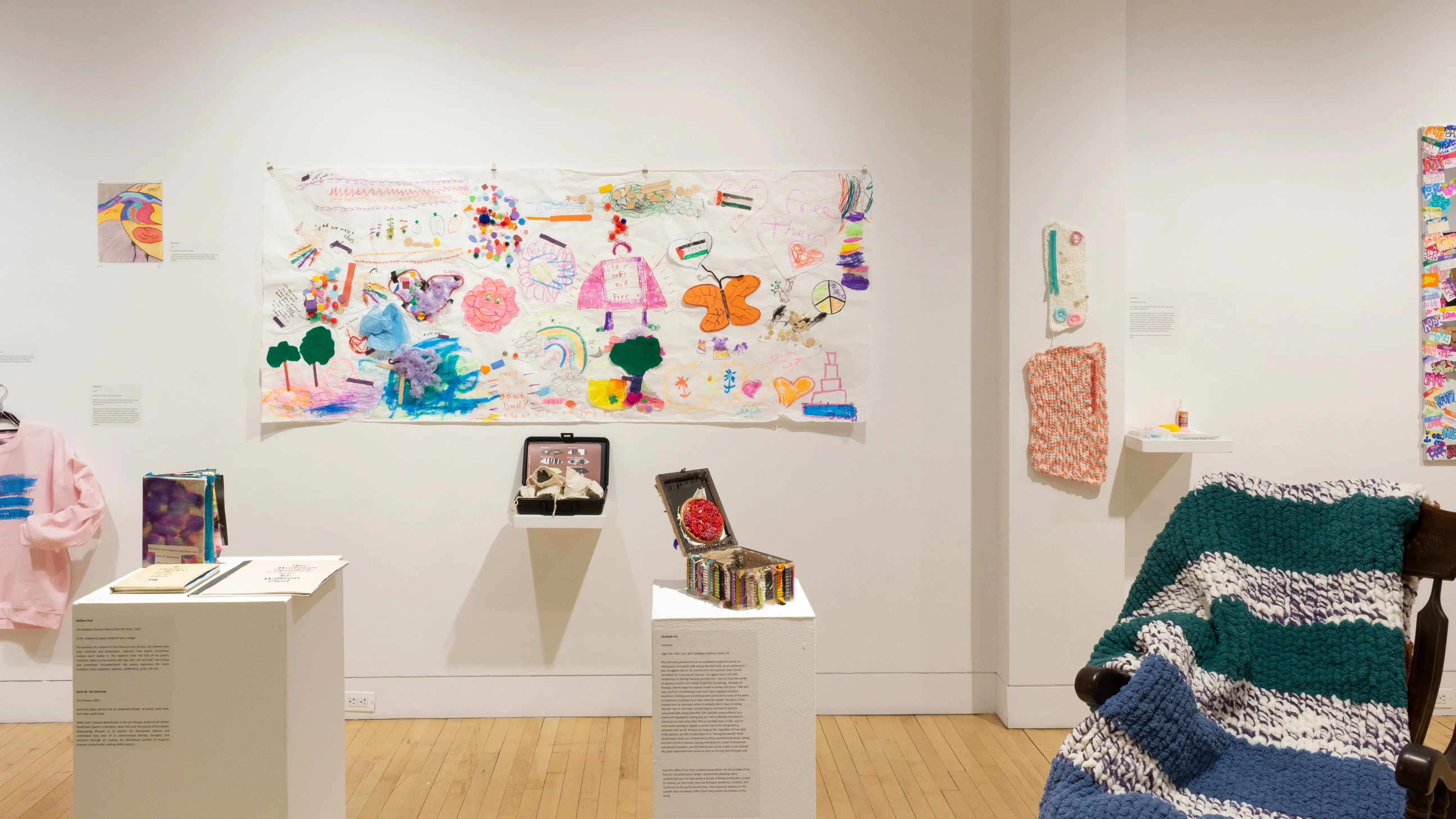 A long drawing on paper hangs on a wall above small sculptures on pedestals and shelves. Textile works hang on the wall to the left and right of the drawing.