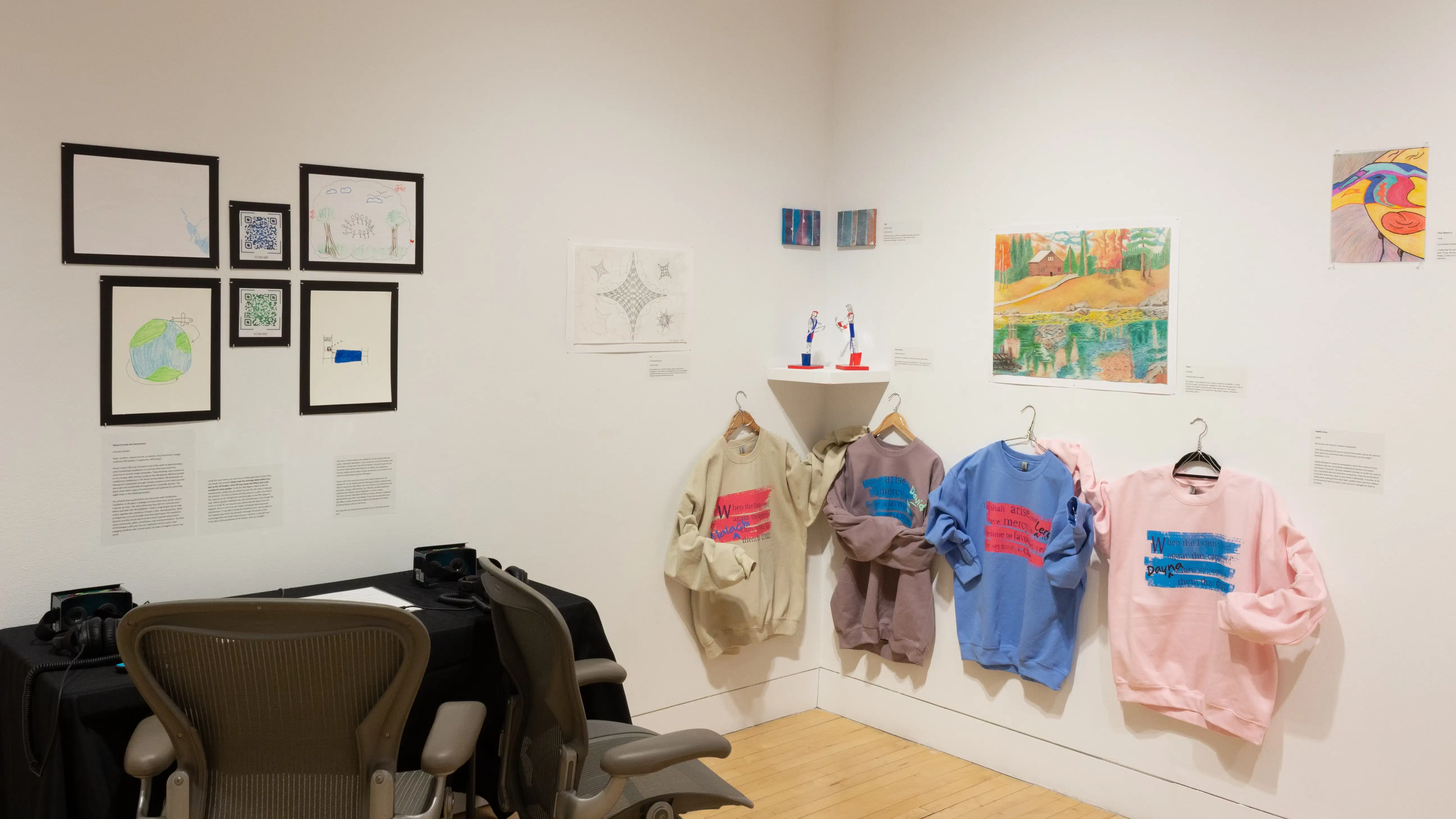 In one corner of the gallery, there are two office chairs facing a table and several framed works hanging above it. To the right, a row of sweatshirts are printed with pink and blue text. Around them hang various drawings and small sculptures.