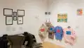 In one corner of the gallery, there are two office chairs facing a table and several framed works hanging above it. To the right, a row of sweatshirts are printed with pink and blue text. Around them hang various drawings and small sculptures.