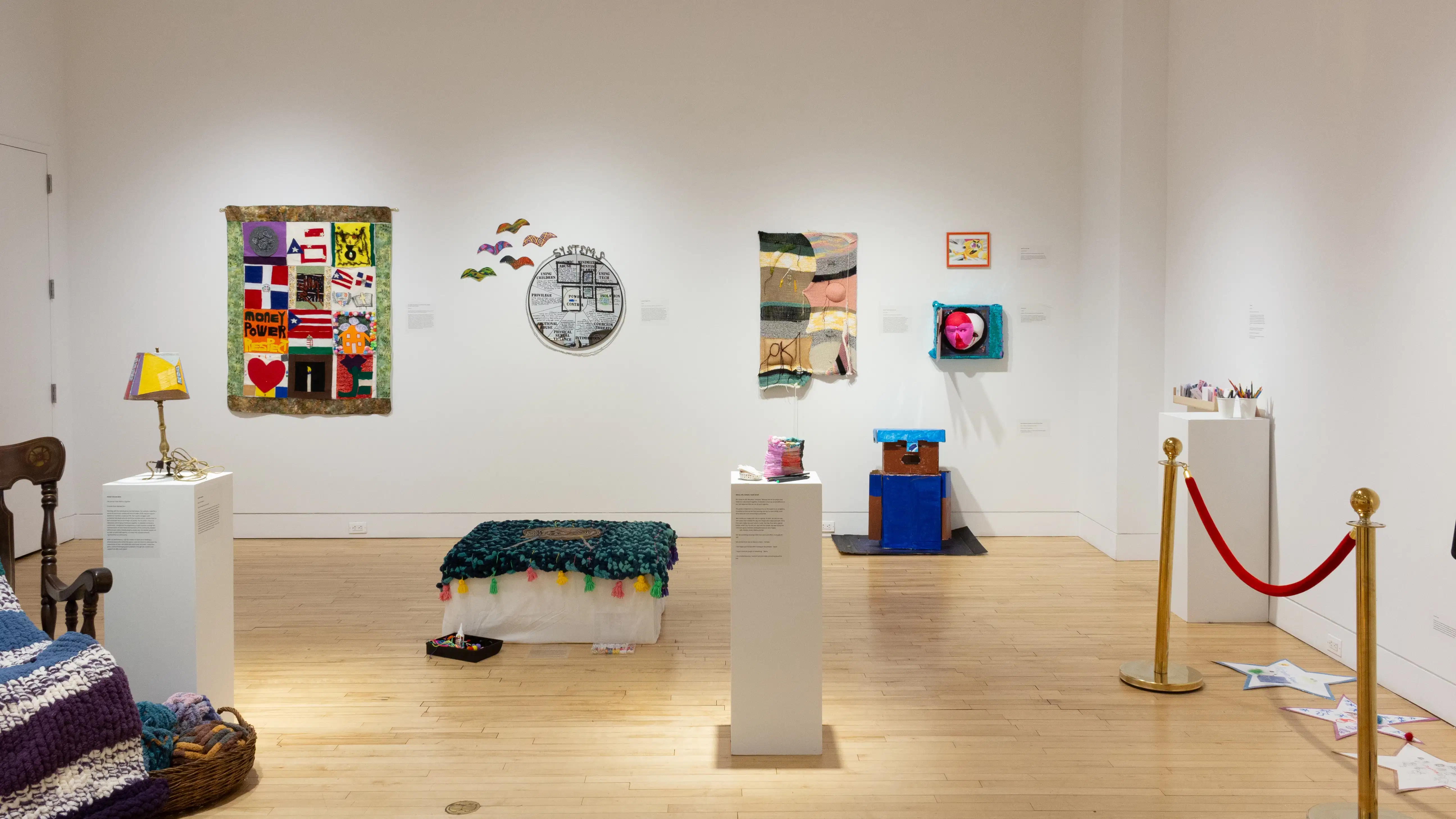 A view of the gallery shows the rocking chair, quilts, ottoman, lamp, paper stars, and stanchion, in addition to other works of various mediums.