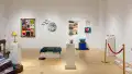 A view of the gallery shows the rocking chair, quilts, ottoman, lamp, paper stars, and stanchion, in addition to other works of various mediums.