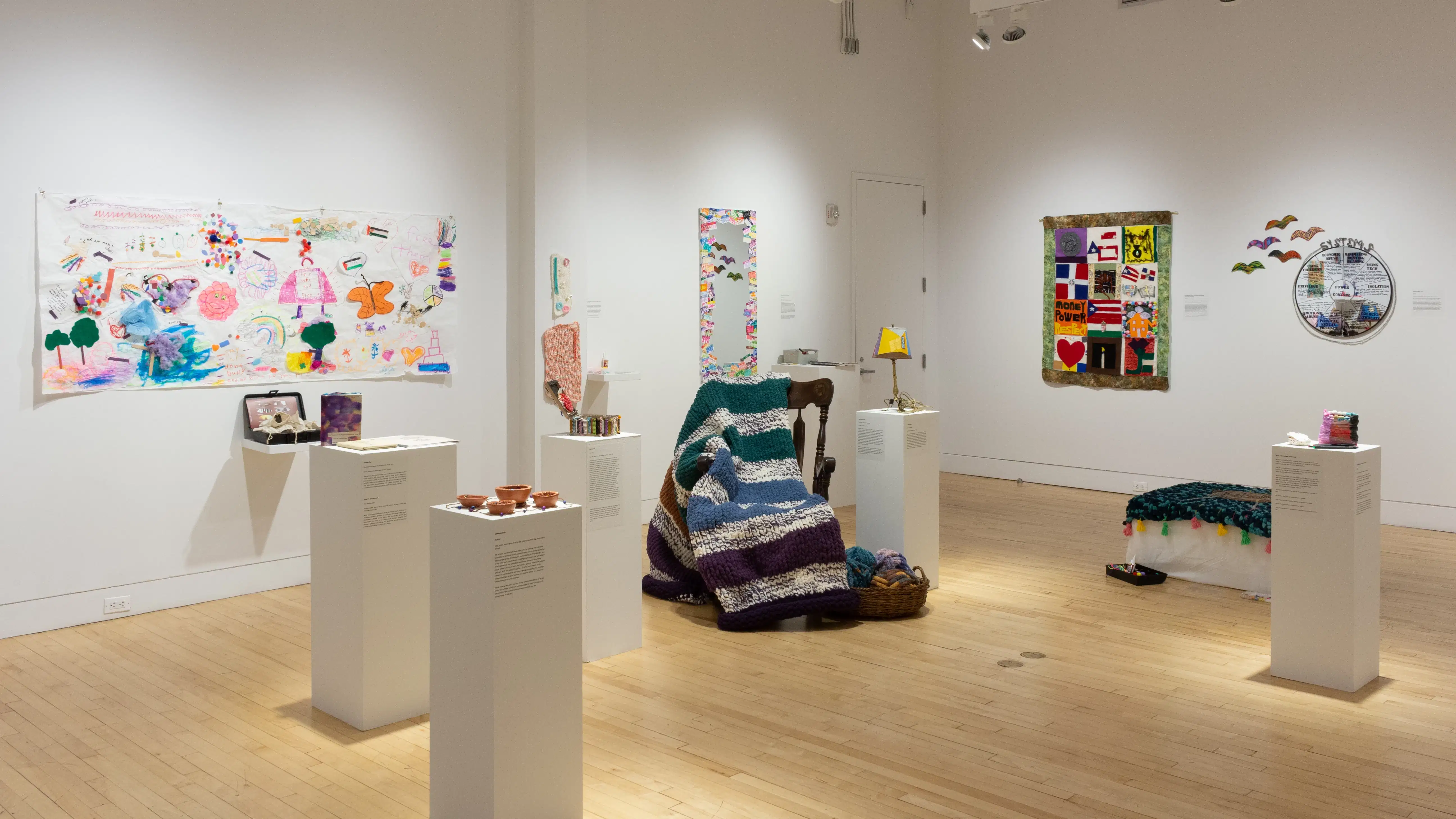 Pedestals holding small sculptures surround a rocking chair and blanket, which sit in the center of the room. Colorful drawings and textile works hang on the walls behind them.