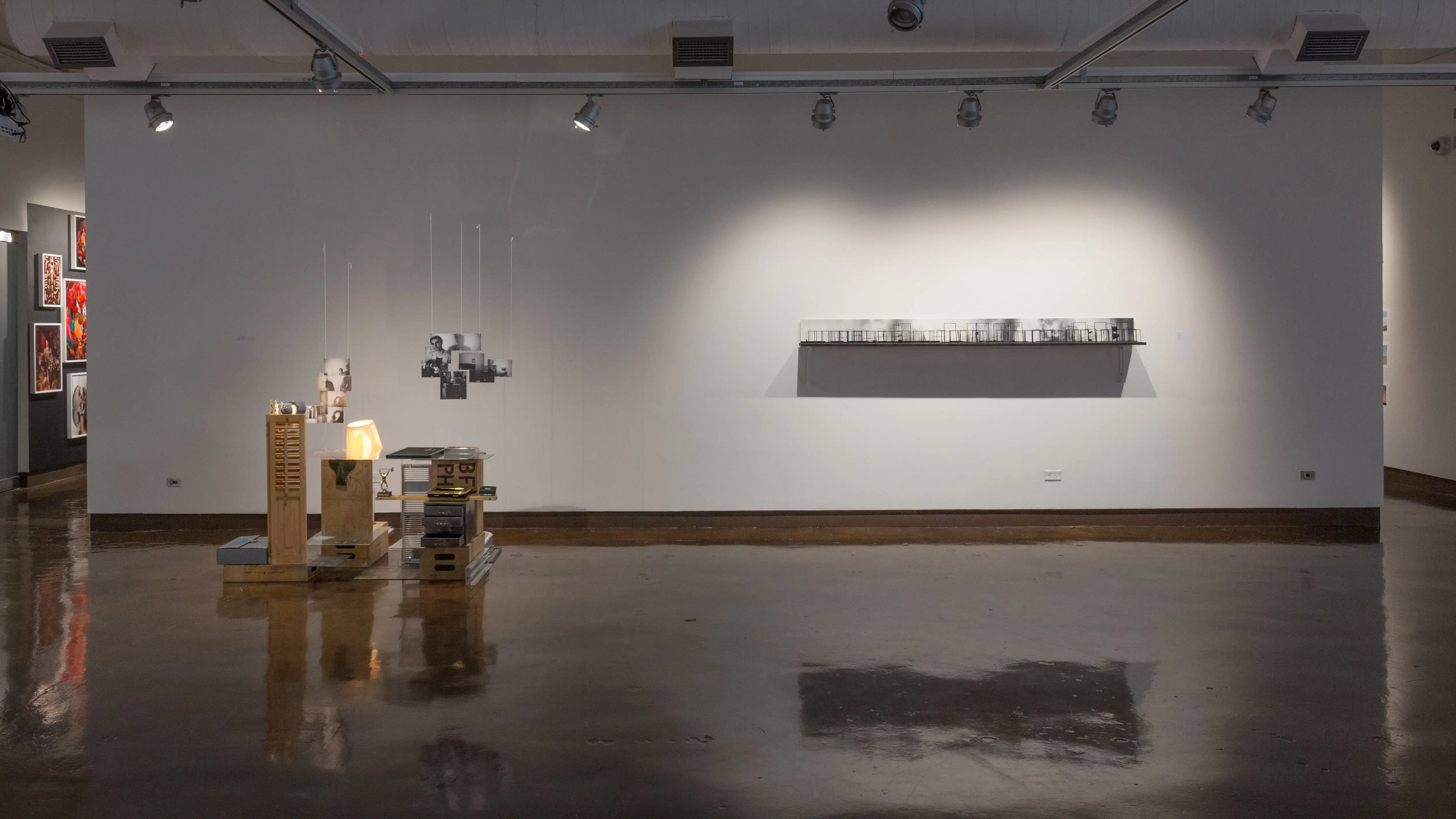 Still in gallery one, an installation featuring a lamp, a box fan, and wooden crates sits next to a shelf holding photos and small black brackets.