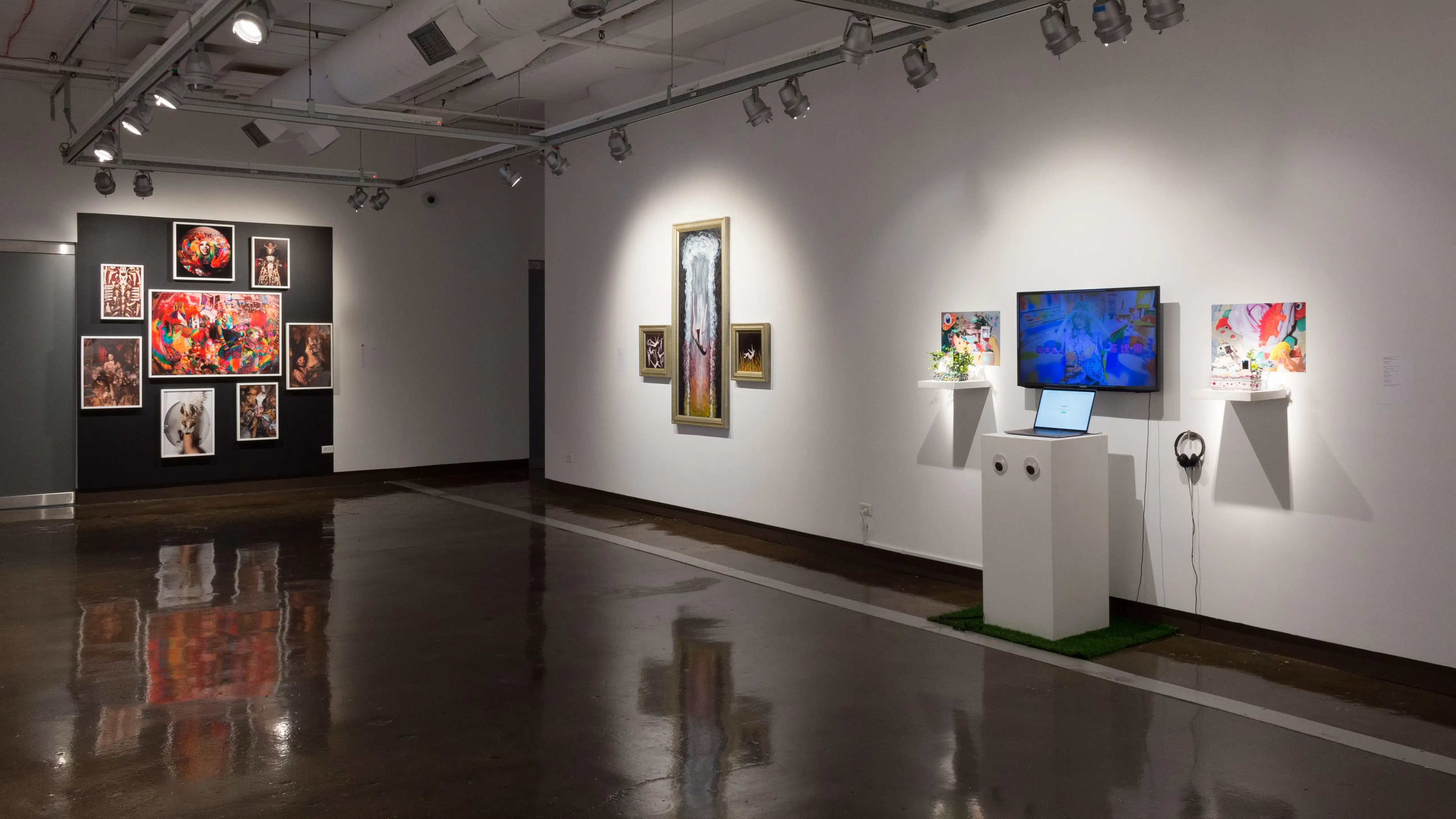 In gallery two, a black wall at the far end holds several brightly colored framed photos, arranged in a circular format. On a long white wall to the right, three gold-framed works of painted photographs hang beside a monitor flanked by prints, sculptures, and a laptop.