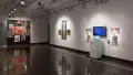 In gallery two, a black wall at the far end holds several brightly colored framed photos, arranged in a circular format. On a long white wall to the right, three gold-framed works of painted photographs hang beside a monitor flanked by prints, sculptures, and a laptop.
