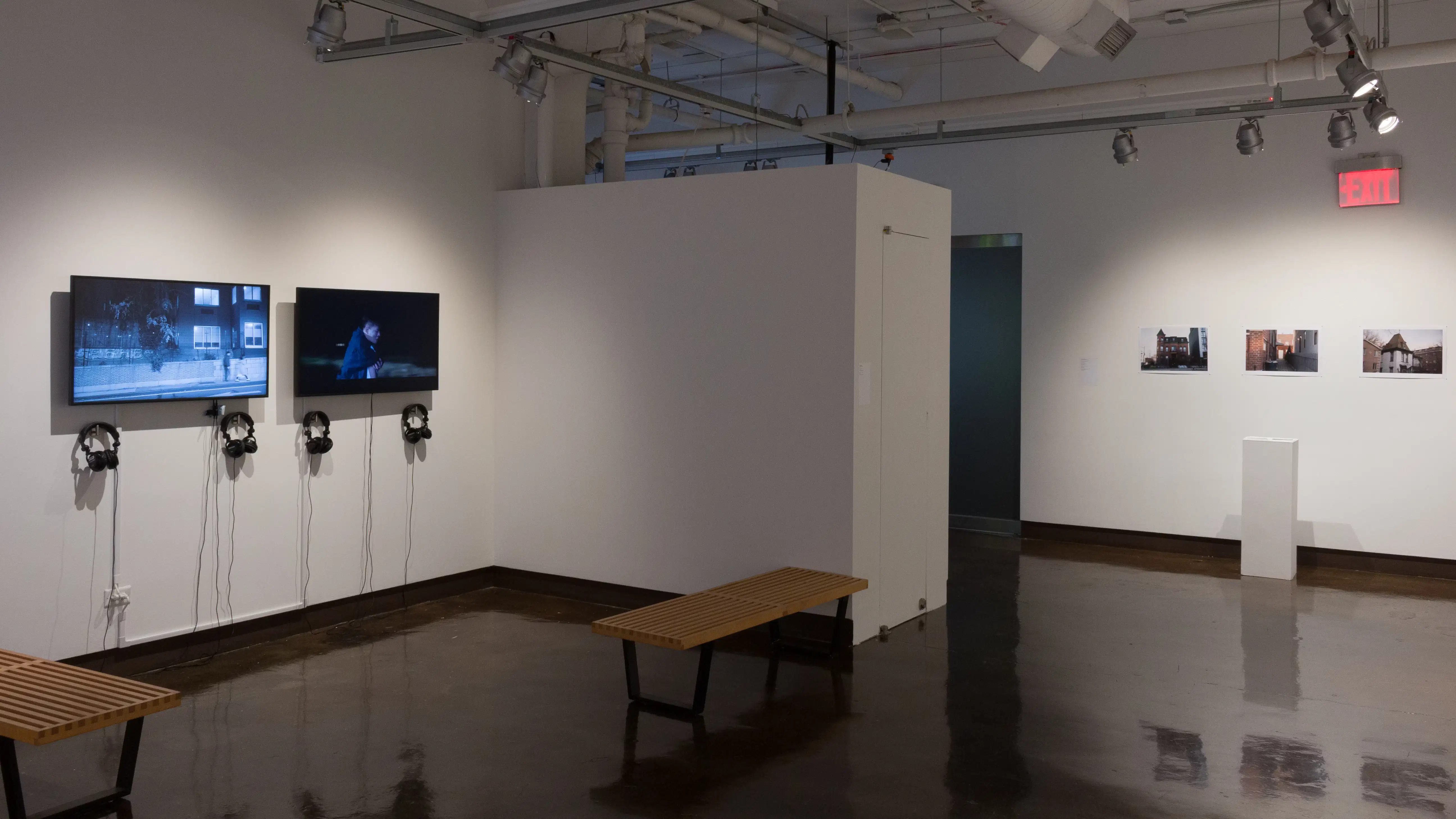 The alcove of gallery four holds monitors displaying different video works, along with headphones to listen to them. On a far wall, a pedestal sits in front of a row of photographs of houses.