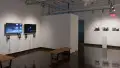 The alcove of gallery four holds monitors displaying different video works, along with headphones to listen to them. On a far wall, a pedestal sits in front of a row of photographs of houses.