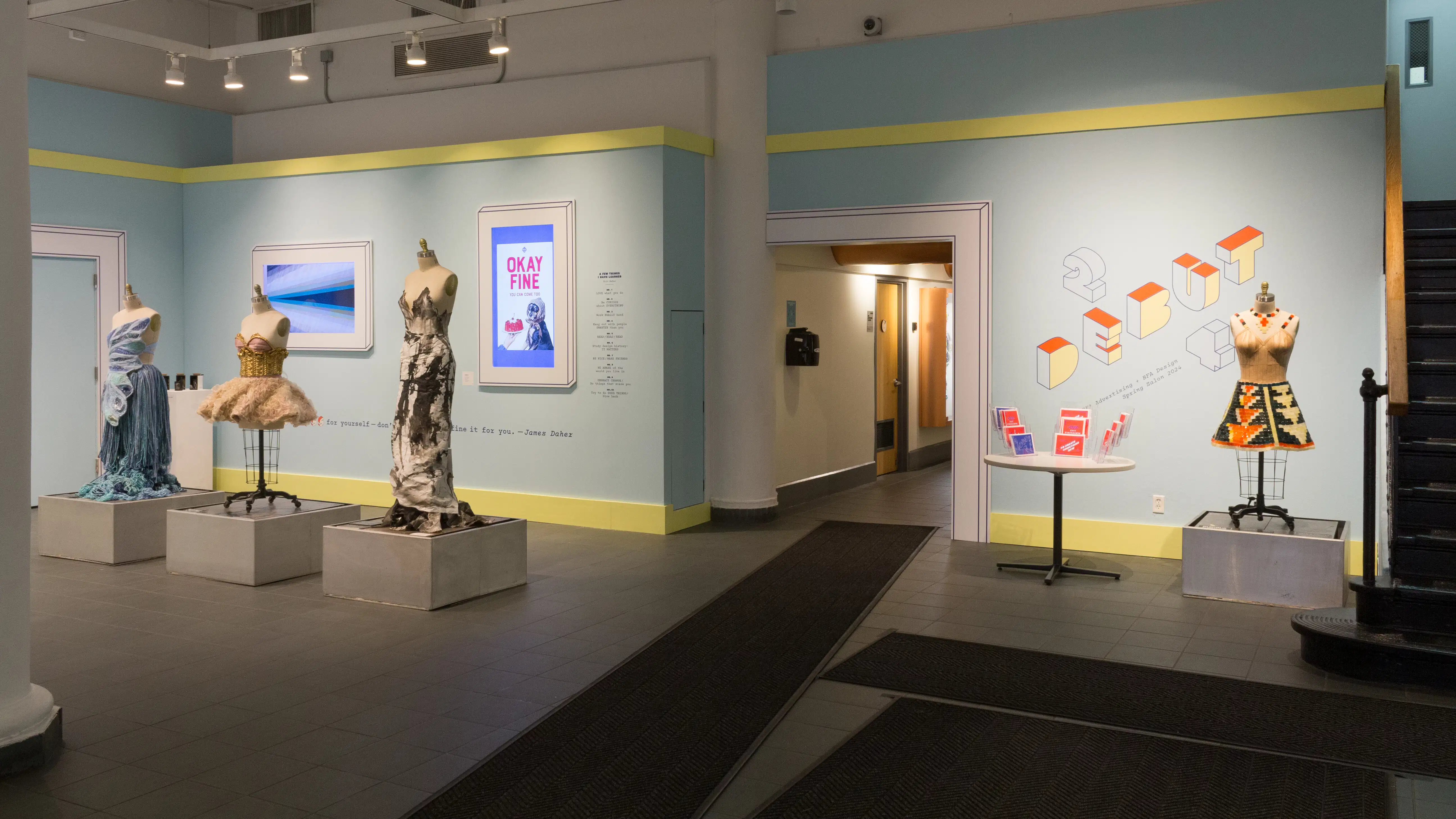 A wide view of the entrance at SVA Gramercy Gallery, showing four dressed mannequins in a row in the center and two screens on the wall. A hallway in the center of the frame leads backwards.