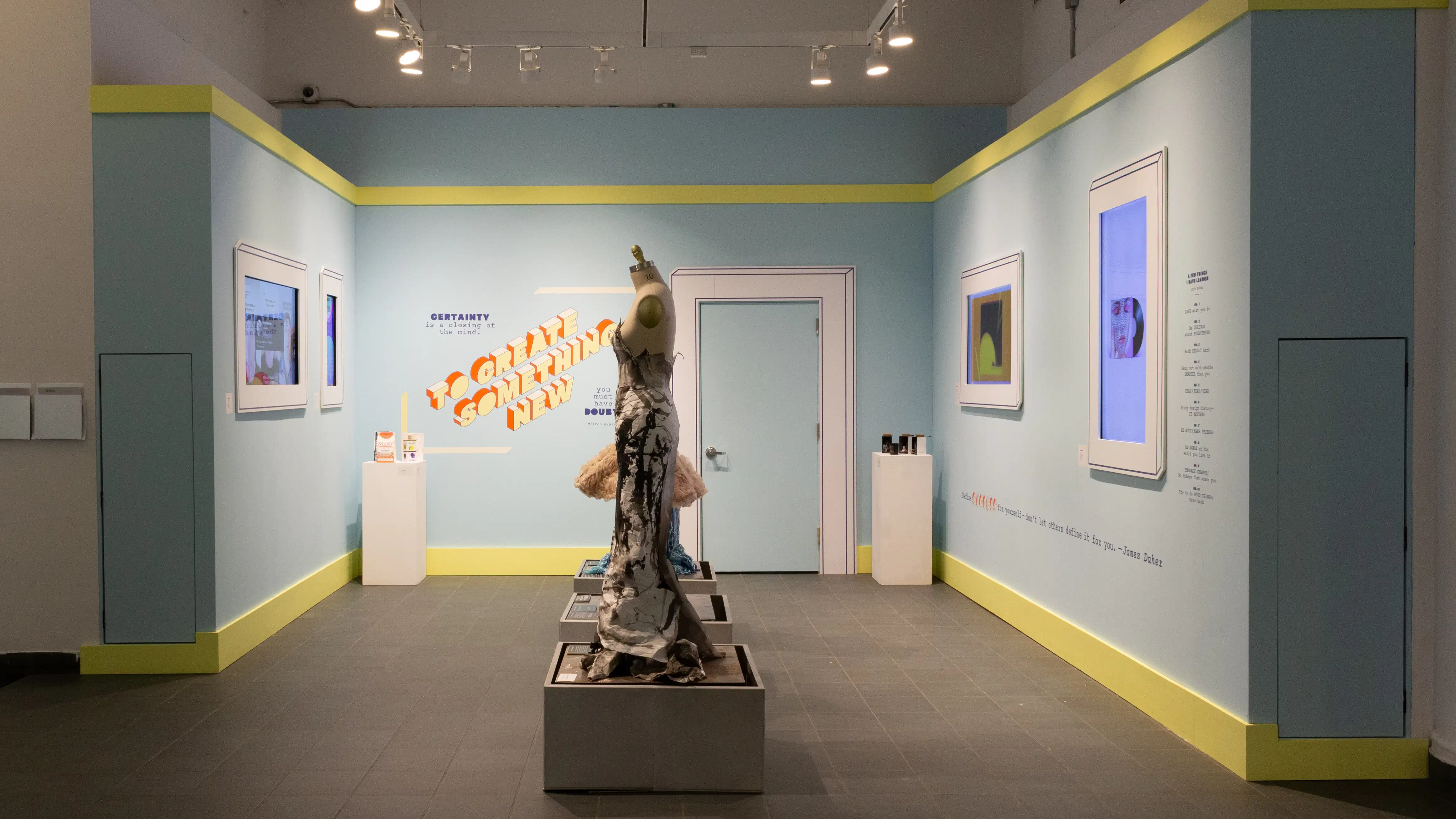 View of the interior alcove of the lobby gallery. There are mannequins in a row down the middle, and two screens each on the opposing walls. The walls are painted blue and the words “To create something new” in 3D type are on the back wall.