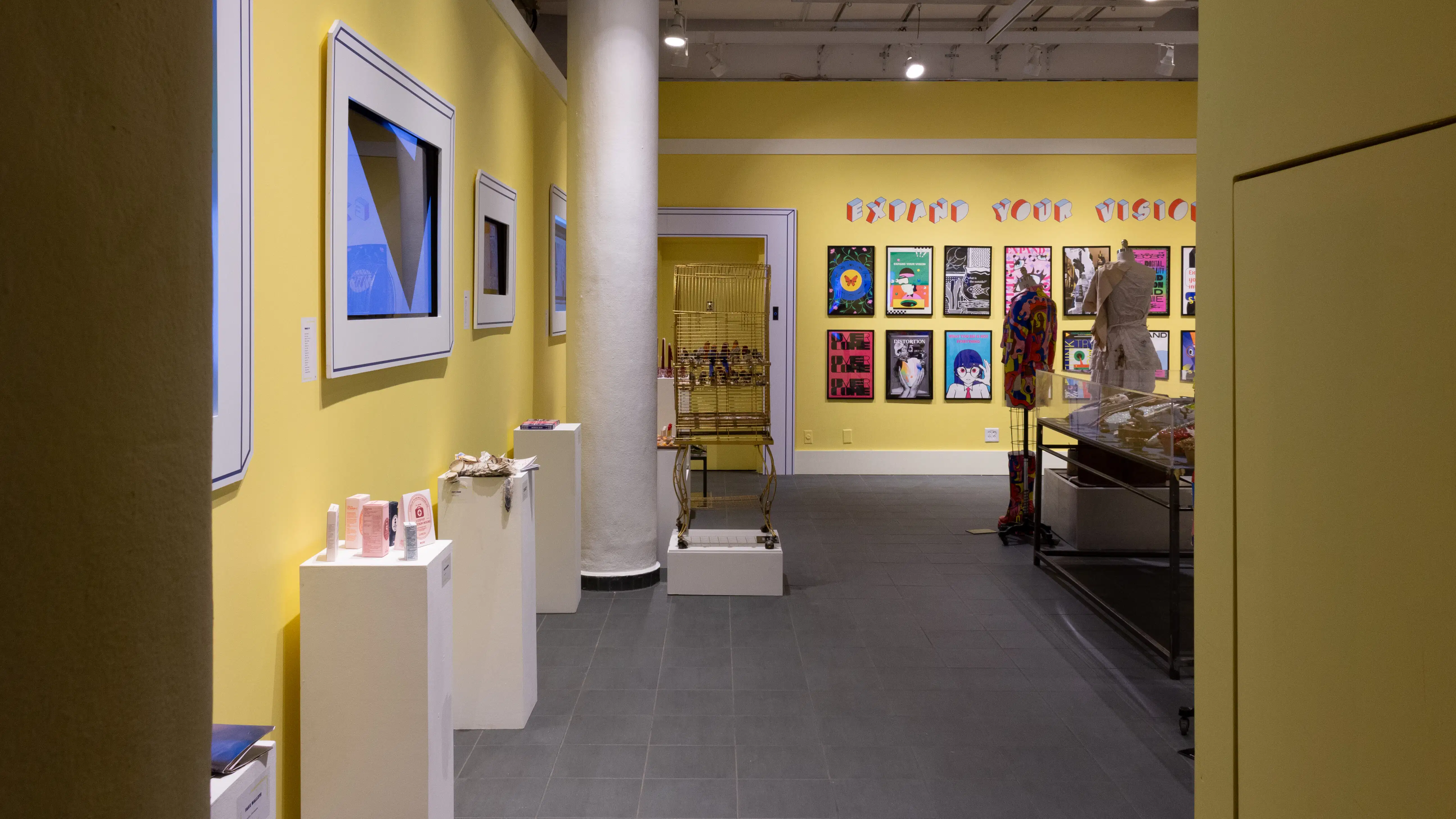 View into the back gallery from the small hallway. The walls are painted yellow and there are several pedestals and a large vitrine in view.