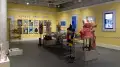 Wide view of gallery two at the Gramercy location. A large plexi vitrine on metal legs can be seen in the middle of the room, filled with painted and adorned ukuleles. Mannequins surround the vitrine and posters and monitors line the walls.