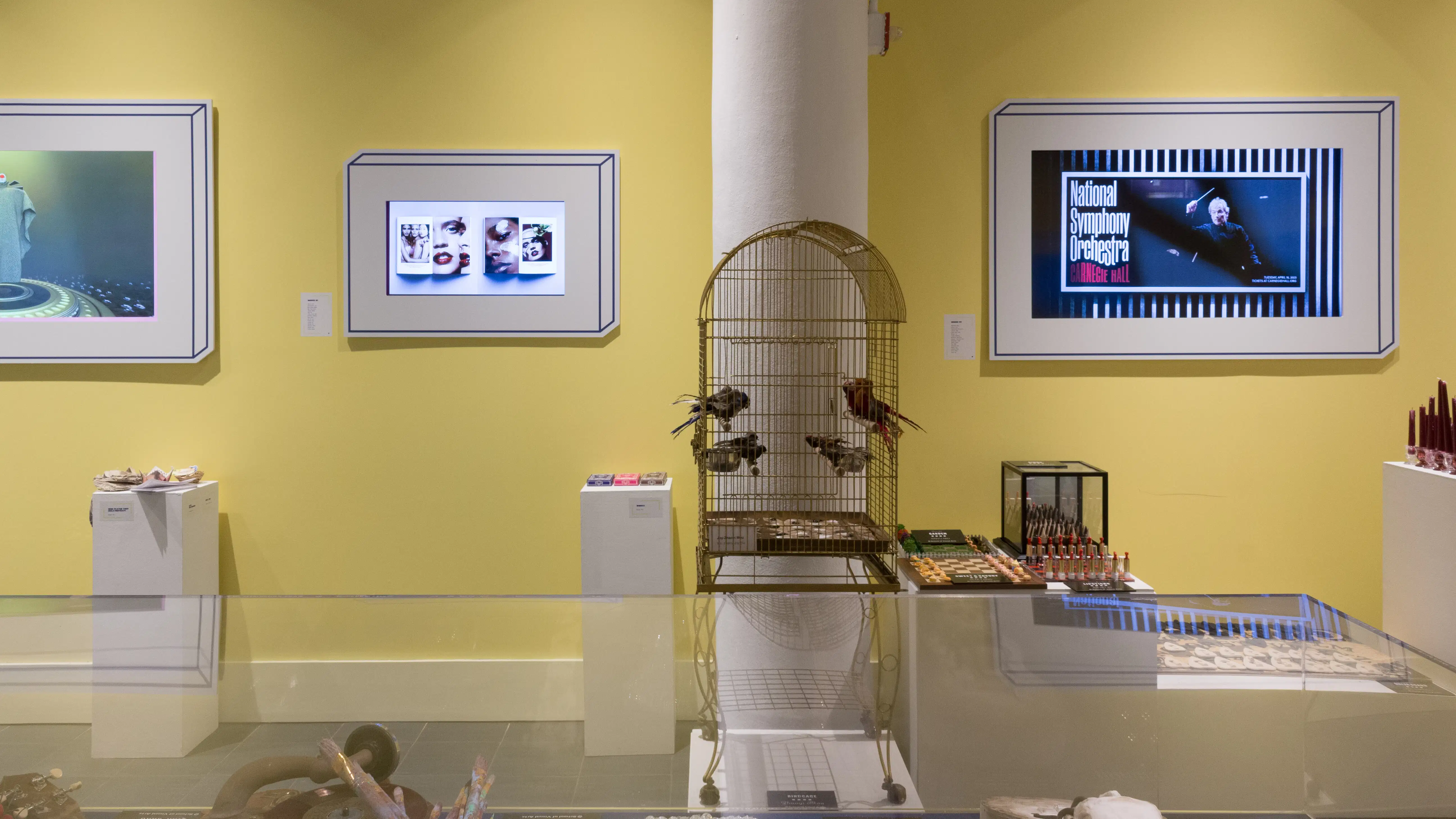 Another view of the left wall upon entering the Gramercy Gallery. A birdcage sits on a pedestal against a column in the center; two monitors can be seen on the left and one on the right.