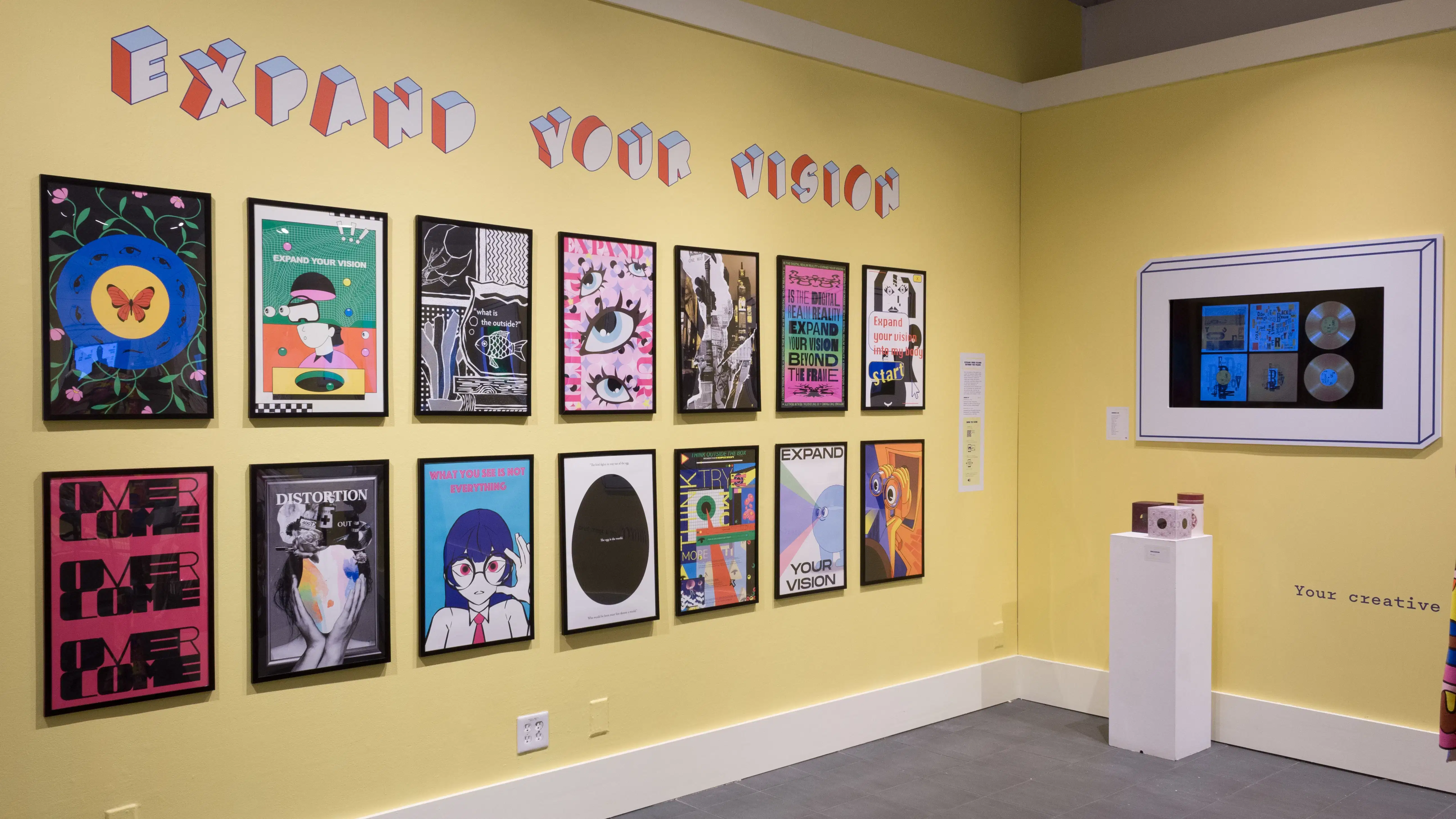 Angled view of the back wall. Fourteen posters line the back wall, and a pedestal sits against a wall with a monitor opposing them.