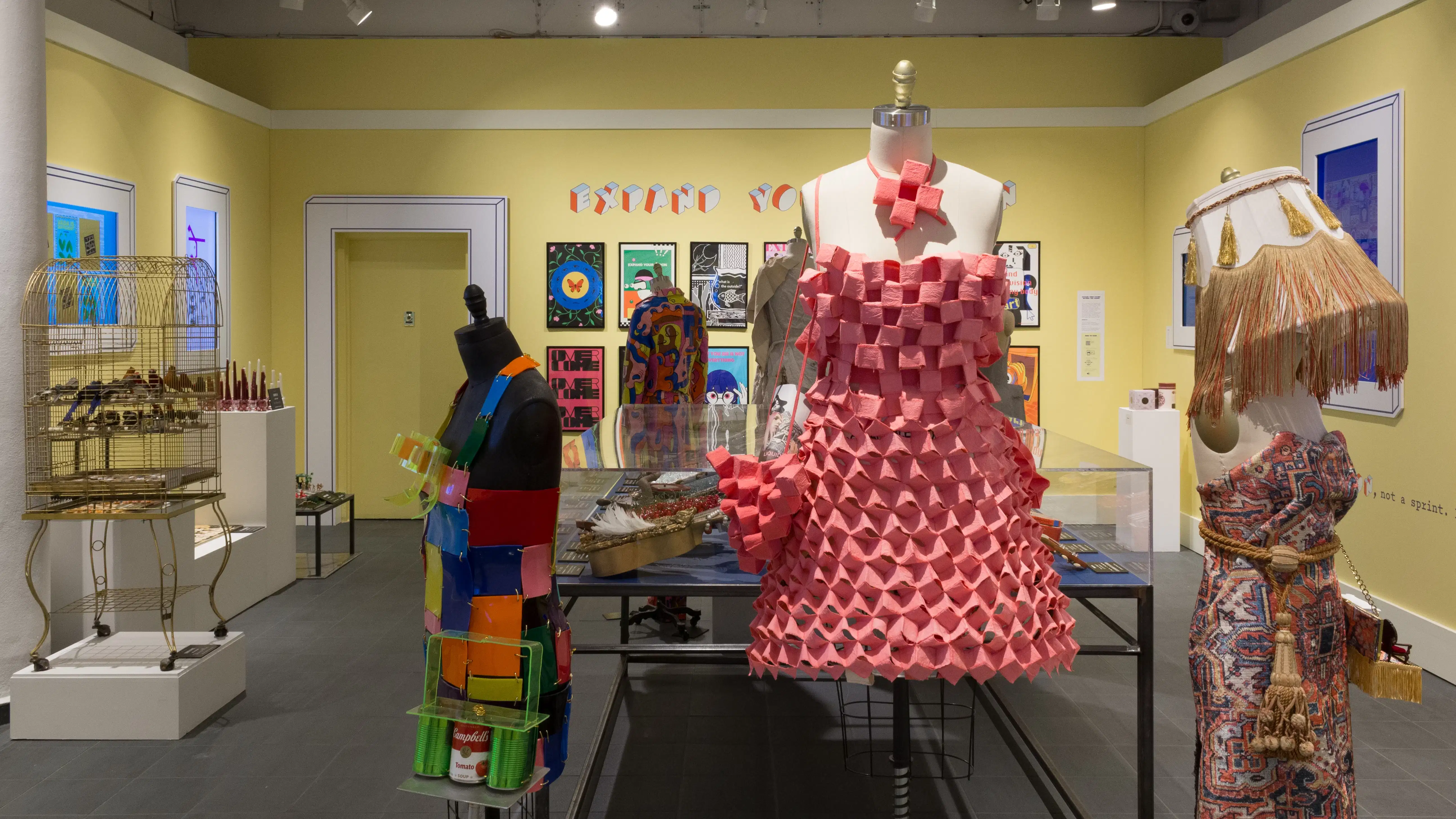 Straight on view of the room, with a mannequin in a pink dress in the middle foreground, a mannequin with a lampshade hat on the right, and a mannequin in a colorful dress on the left, with the vitrine in the background and all of the other installations throughout the room.