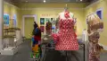 Straight on view of the room, with a mannequin in a pink dress in the middle foreground, a mannequin with a lampshade hat on the right, and a mannequin in a colorful dress on the left, with the vitrine in the background and all of the other installations throughout the room.