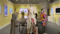 View of the room from the opposite side; a mannequin looms large in the middle foreground with two other mannequins flanking it, the vitrine behind them and monitors displaying colorful images along the walls.
