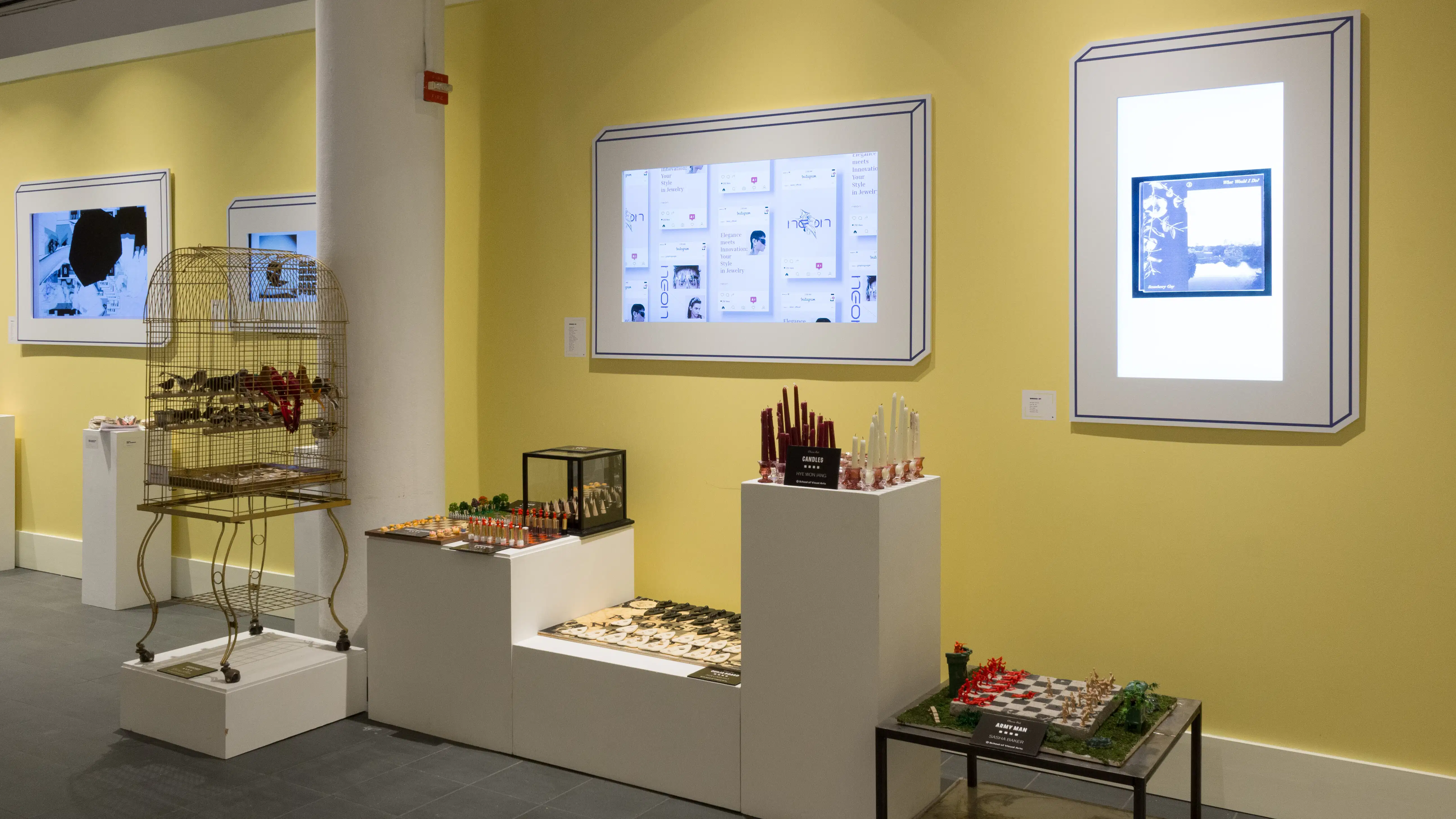 Angled view of the back wall; four installations on pedestals of varying sizes in the foreground, with four brightly-lit monitors along the wall behind them.