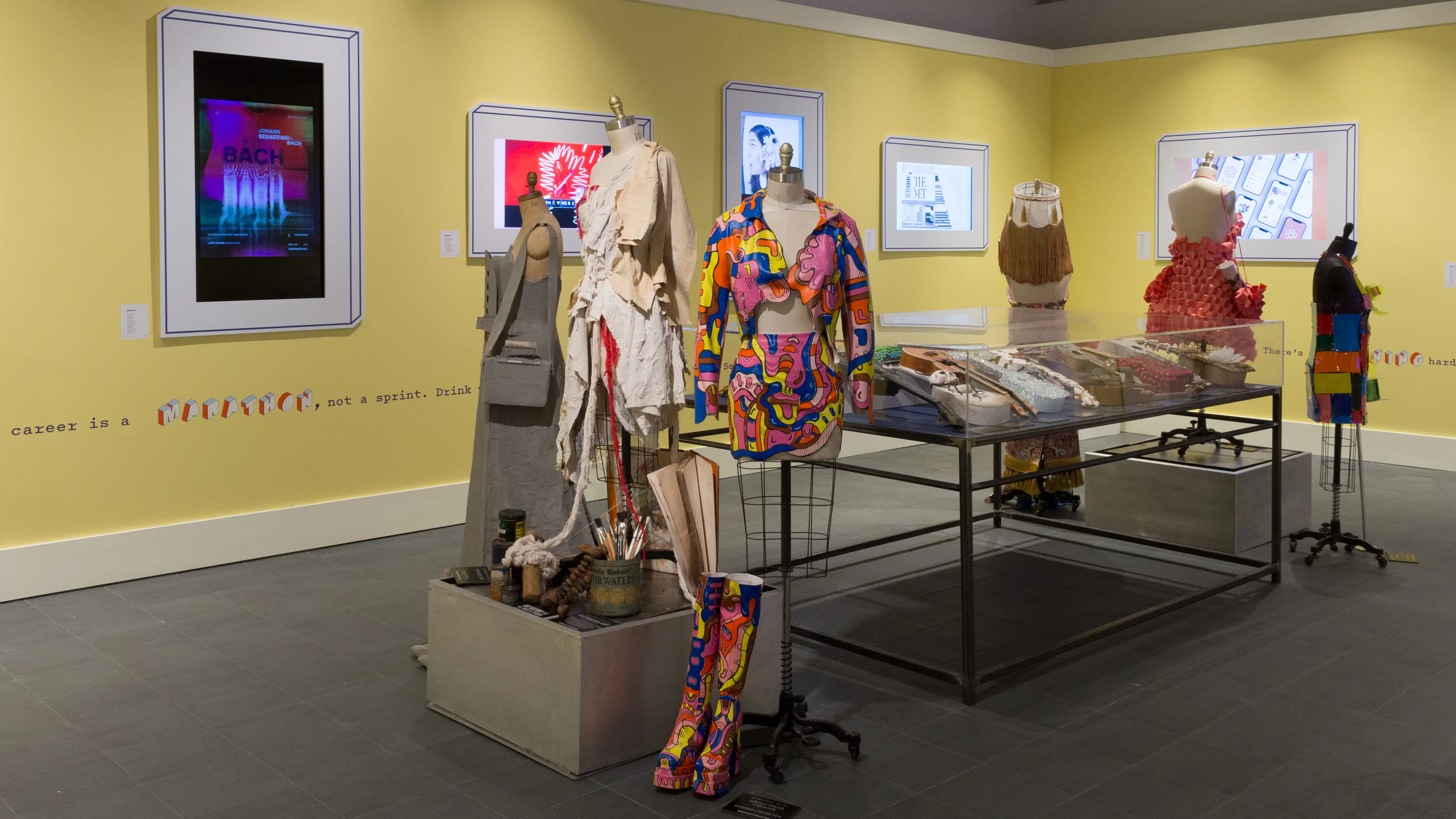 Angled view of the entire room; two mannequin installations with three mannequins each flank a large vitrine on metal legs, and monitors line the walls around them.