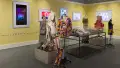 Angled view of the entire room; two mannequin installations with three mannequins each flank a large vitrine on metal legs, and monitors line the walls around them.