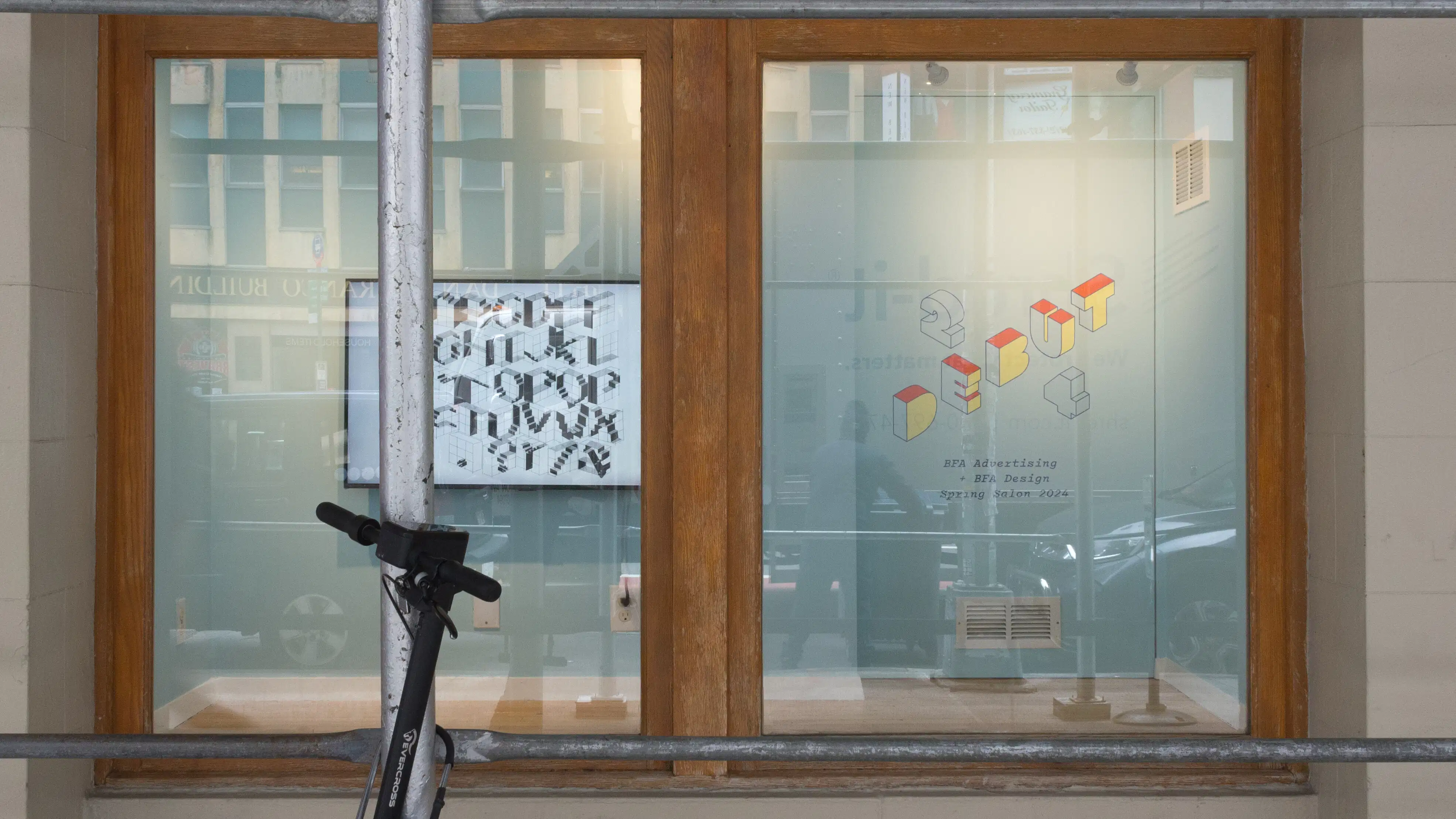 View of the window vitrine from the street; a screen hangs on the wall on the left, and the show title “Debut 24” can be seen in vinyl block letters on the right.
