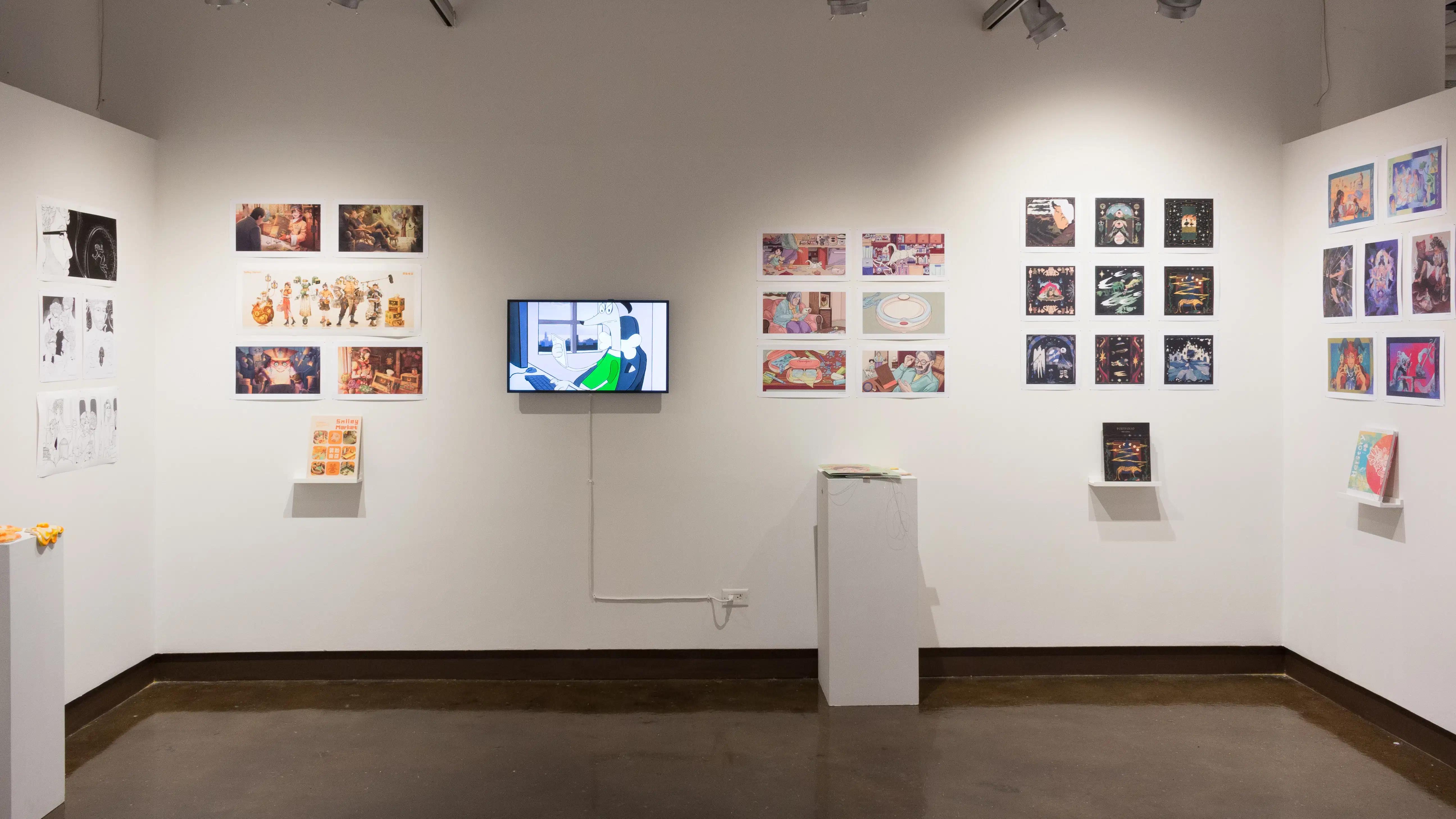 In the alcove of the third gallery space, more student work is displayed in the form of prints and books. However, there is also a monitor playing animations, and a pedestal holding some ceramic sculptures of fried eggs.