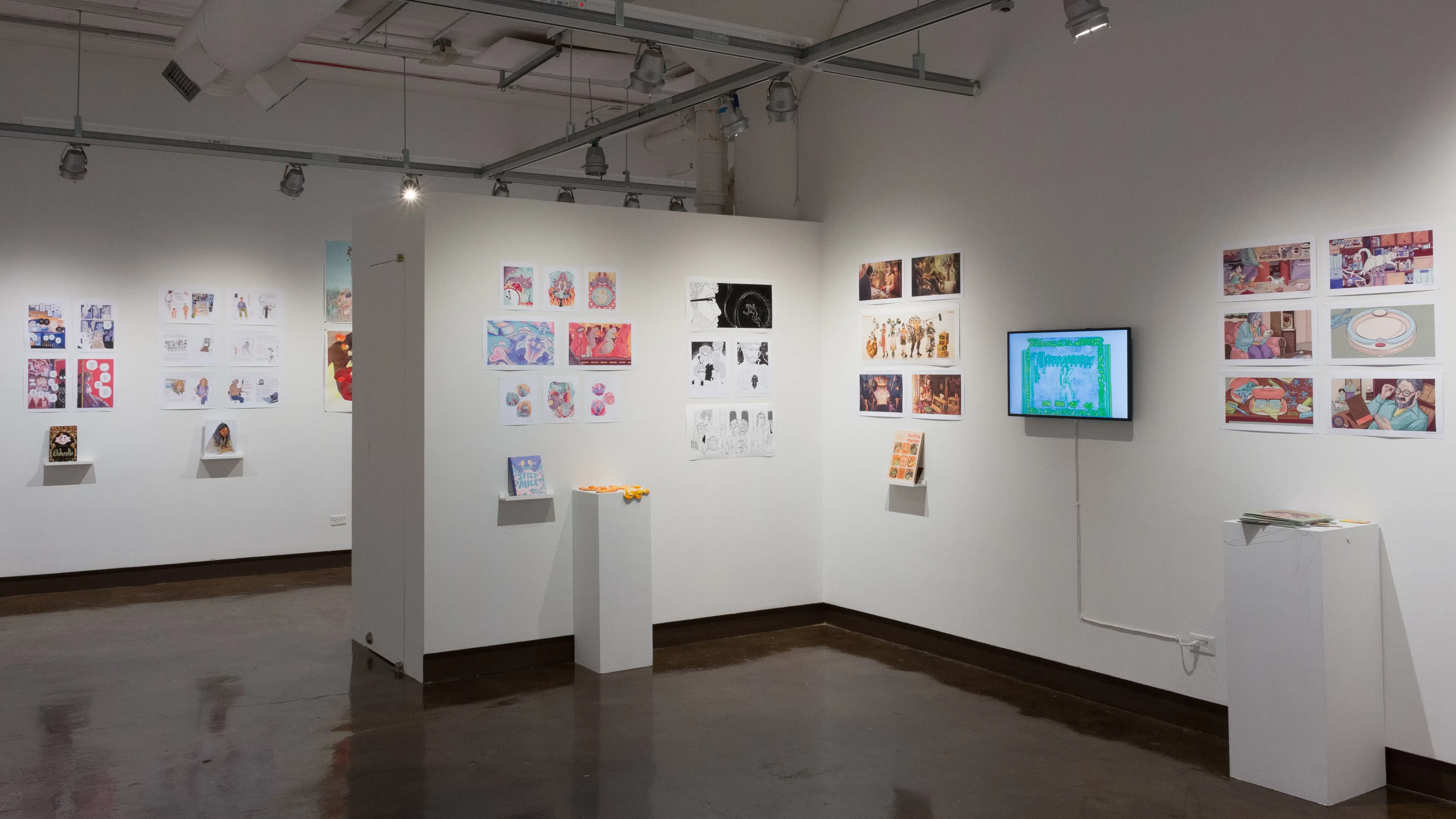 In the third gallery space, the prints hanging on the walls vary in dimension, style, and color. Books sit on shelves beneath the prints and a TV displays online works and animations and beside the TV, a pedestal holding some ceramic sculptures of fried eggs.