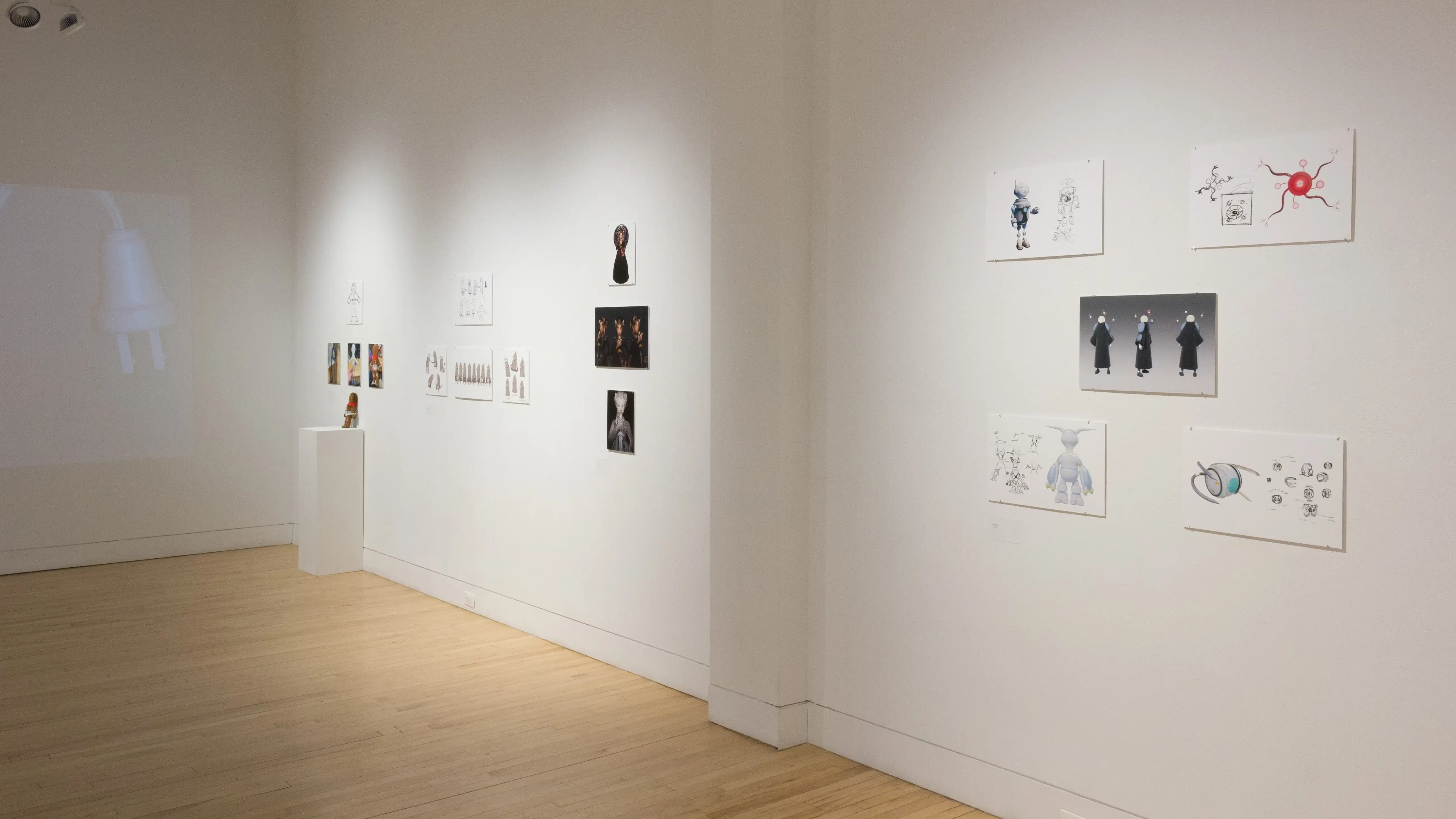 Angled view of five student works, showing printed mockups of animated characters, a 3D character display on a pedestal, and a projection on the back of the wall. 