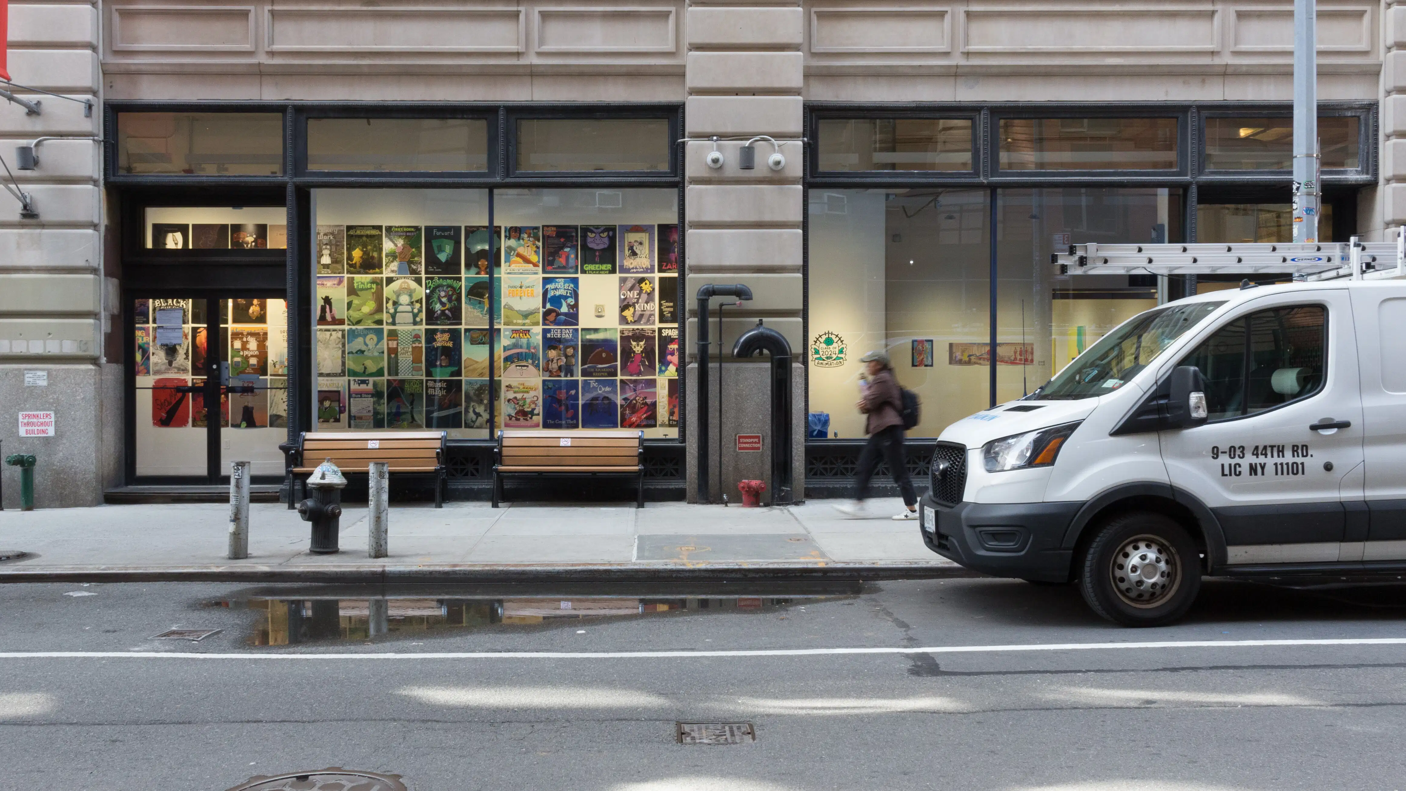 From the street, looking into the windows at the SVA Flatiron Gallery, many posters created by SVA’s BFA Animation Class of 2024 are visible. A person walks by the windows on the sidewalk.