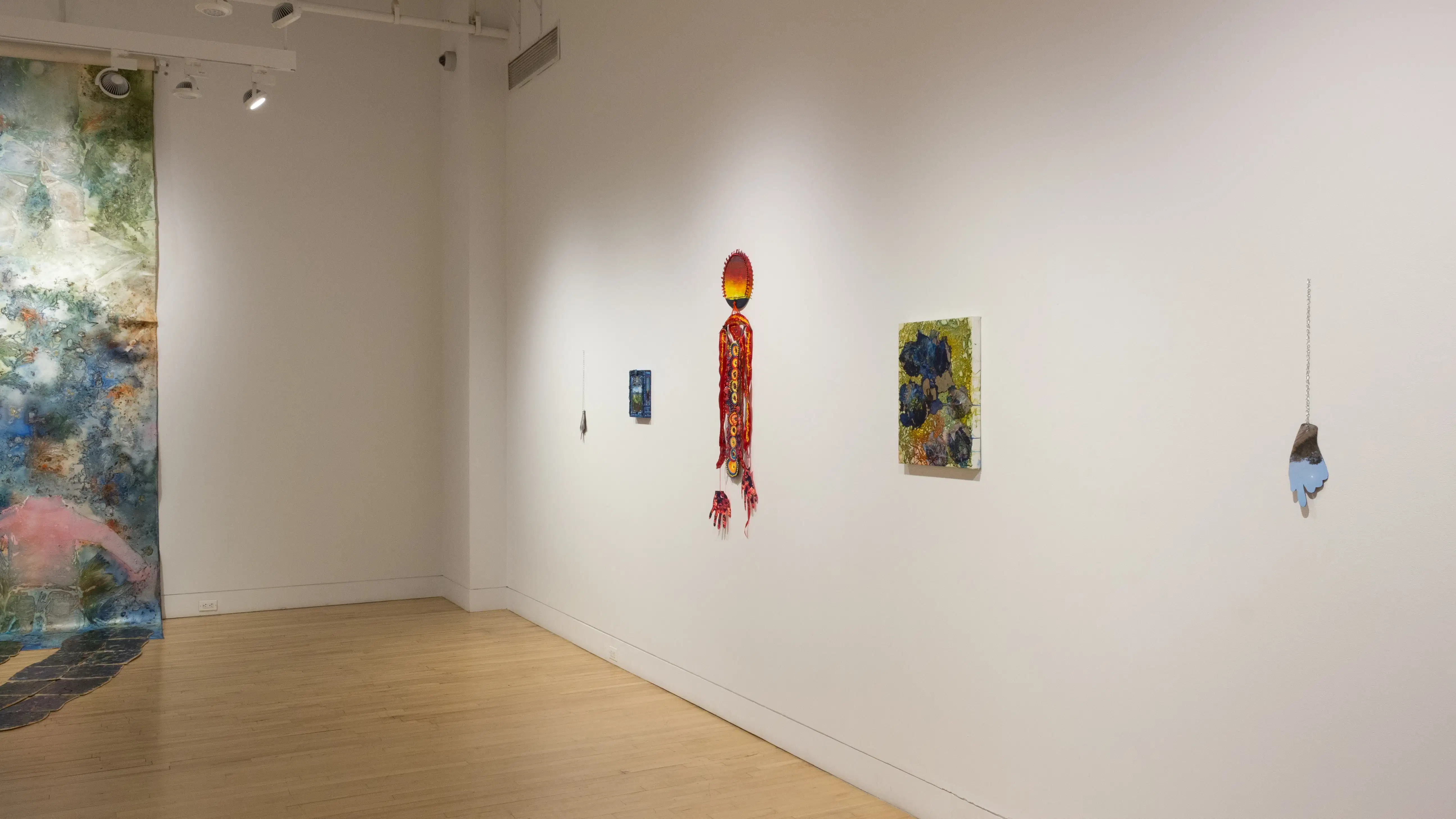 An angled view of the right wall in the SVA Flatiron Gallery, depicting colorful hanging sculpture pieces and paintings. 