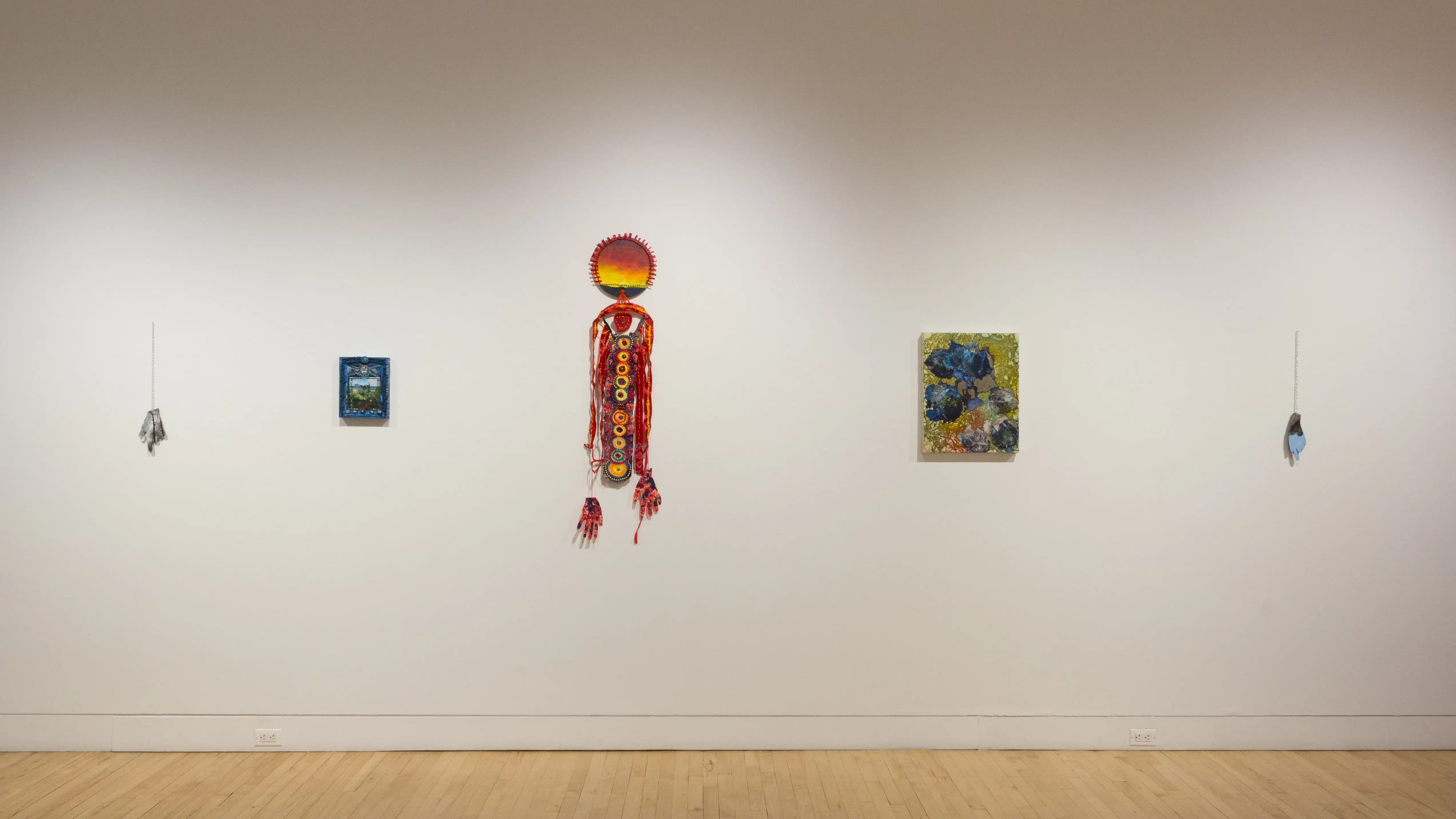 An even view of the right wall in the SVA Flatiron Gallery, displaying colorful hanging sculptures and various sized paintings. 