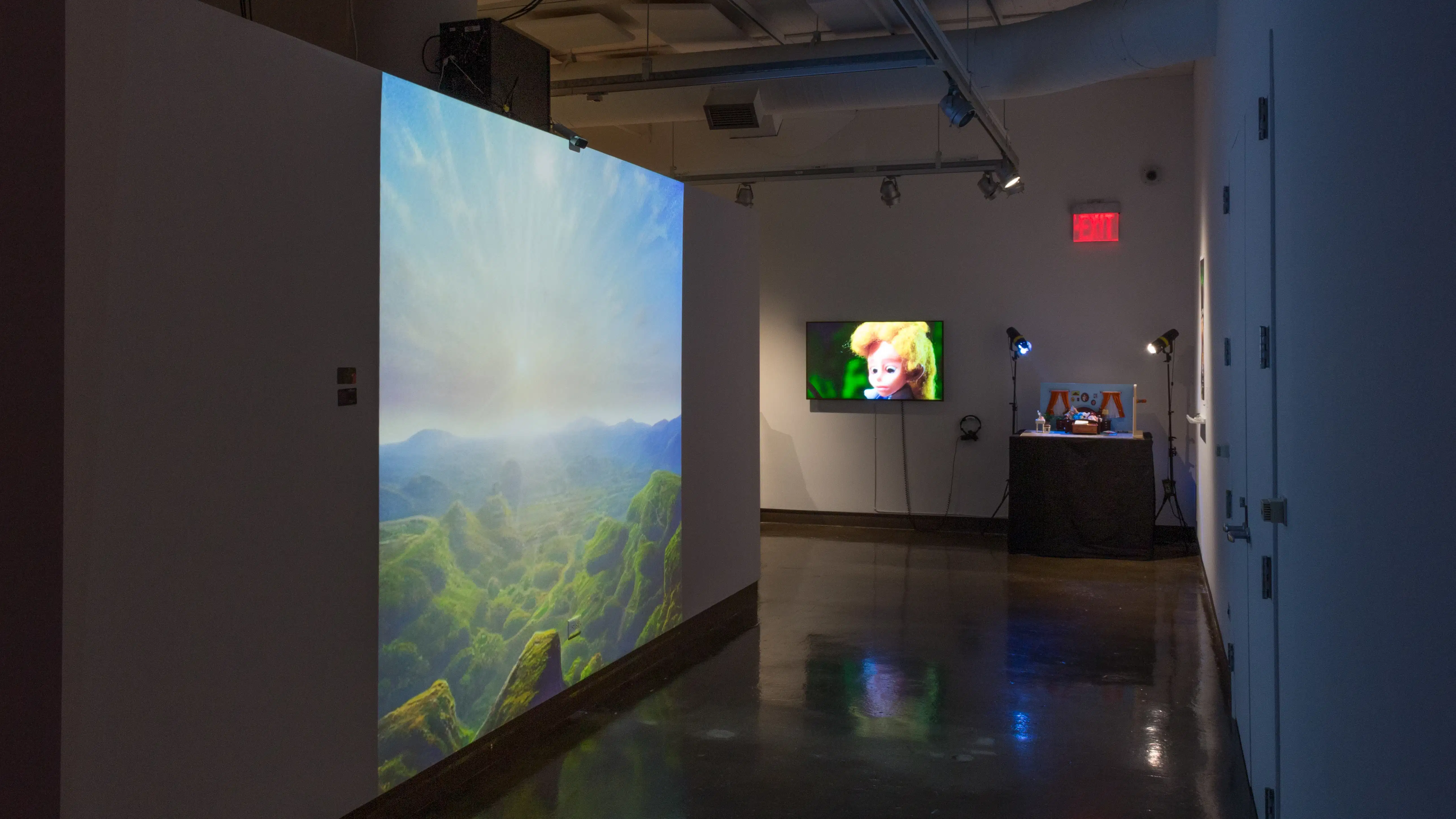 Installation image showing a large projection of a valley to the left, with a monitor and sculptural installation with stand lights in the background.
