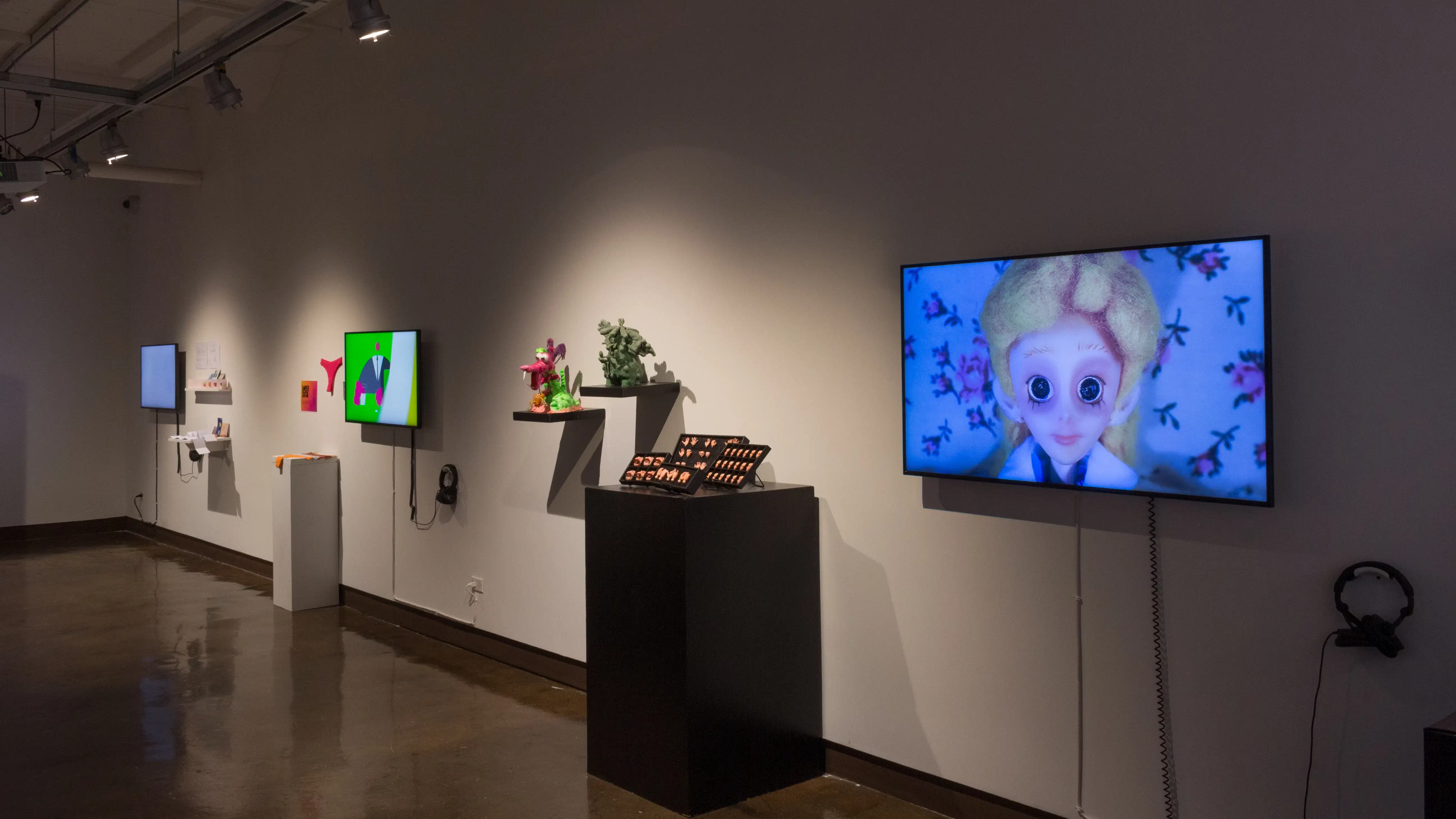 Angled image of a long wall with several monitors, pedestals and shelves with various installations and animated videos.