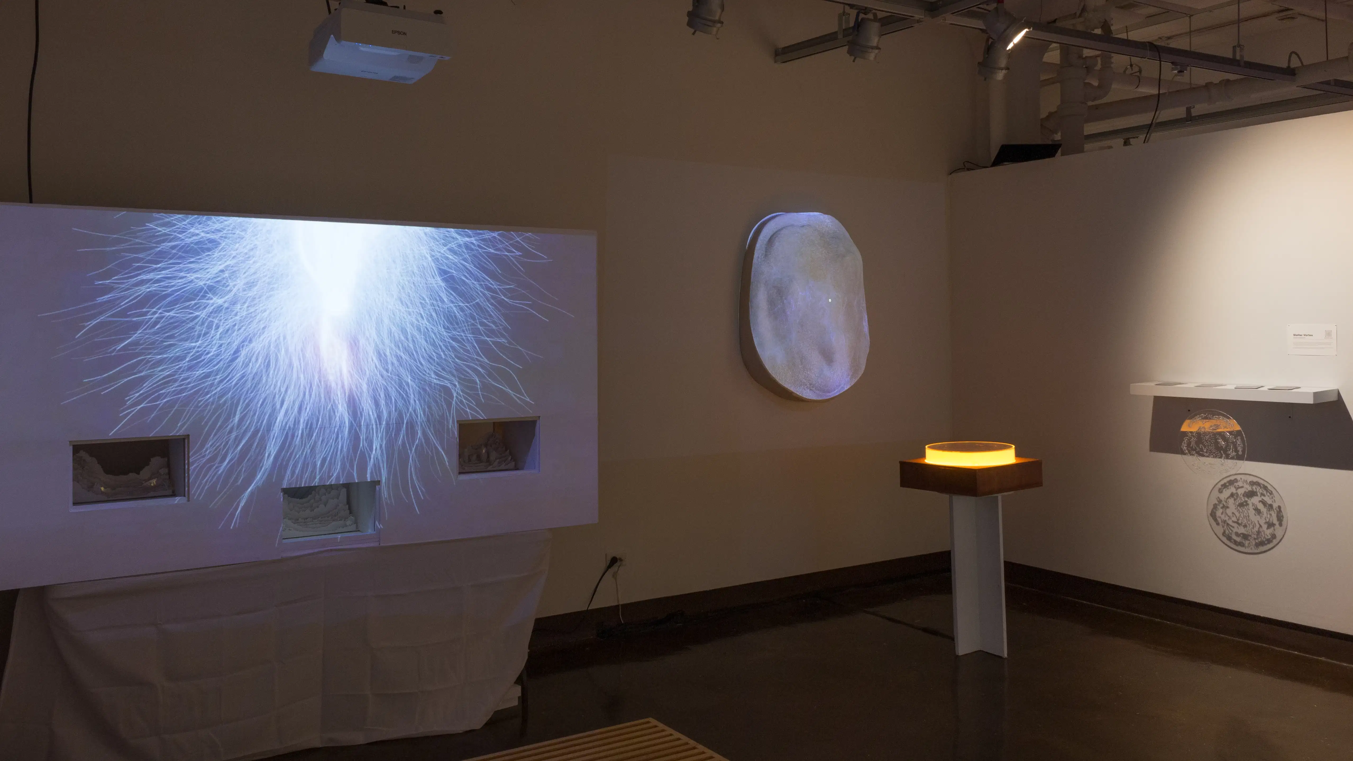 Installation image of an alcove with a large projection of a wavy burst on a false wall with cutouts to the left, and a pedestal and rock projection in the background.