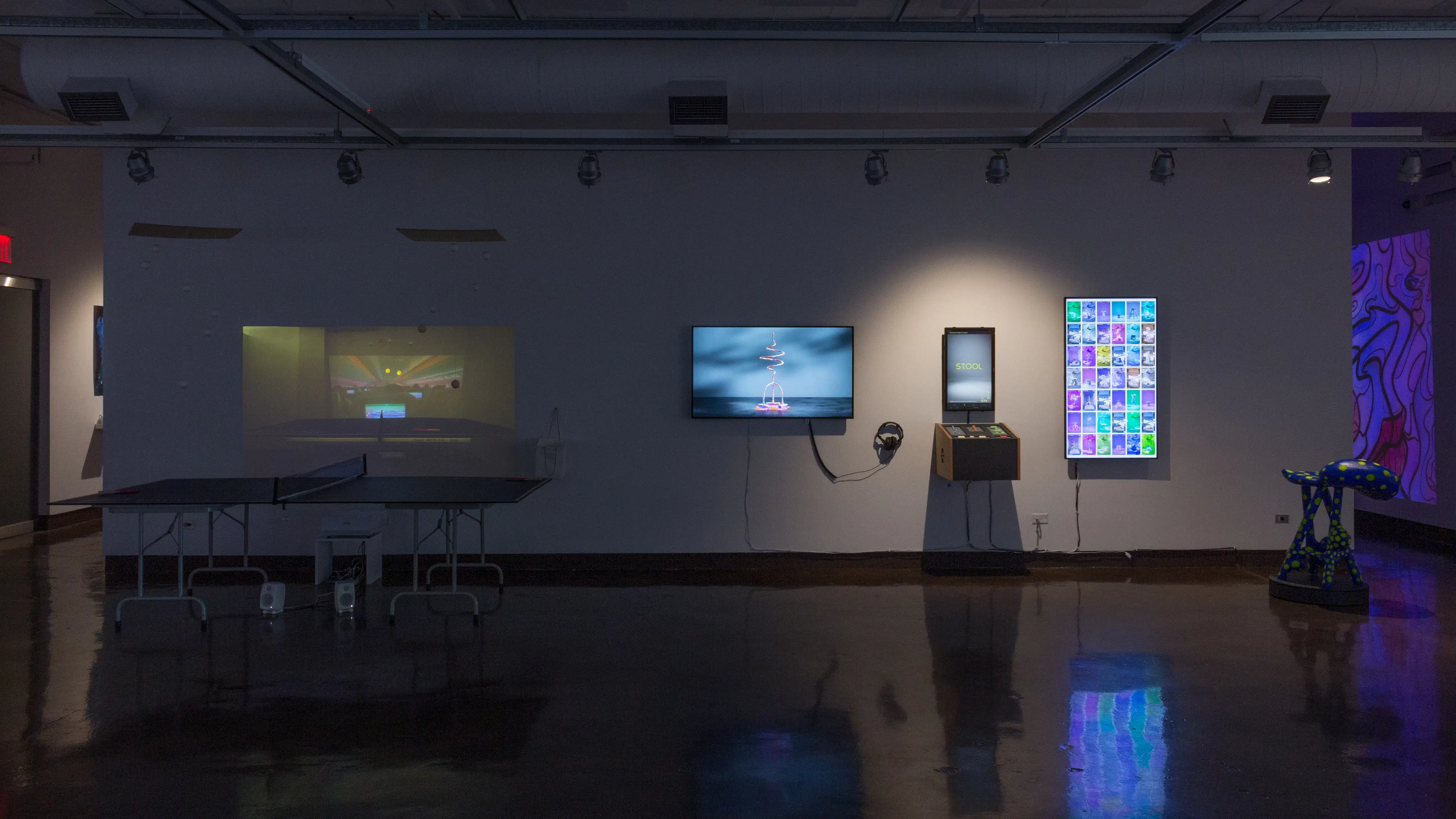 Installation image showing a freestanding wall with a pingpong table in front of a projection on the left, followed by three monitors to the right with an interactive spinning stool.