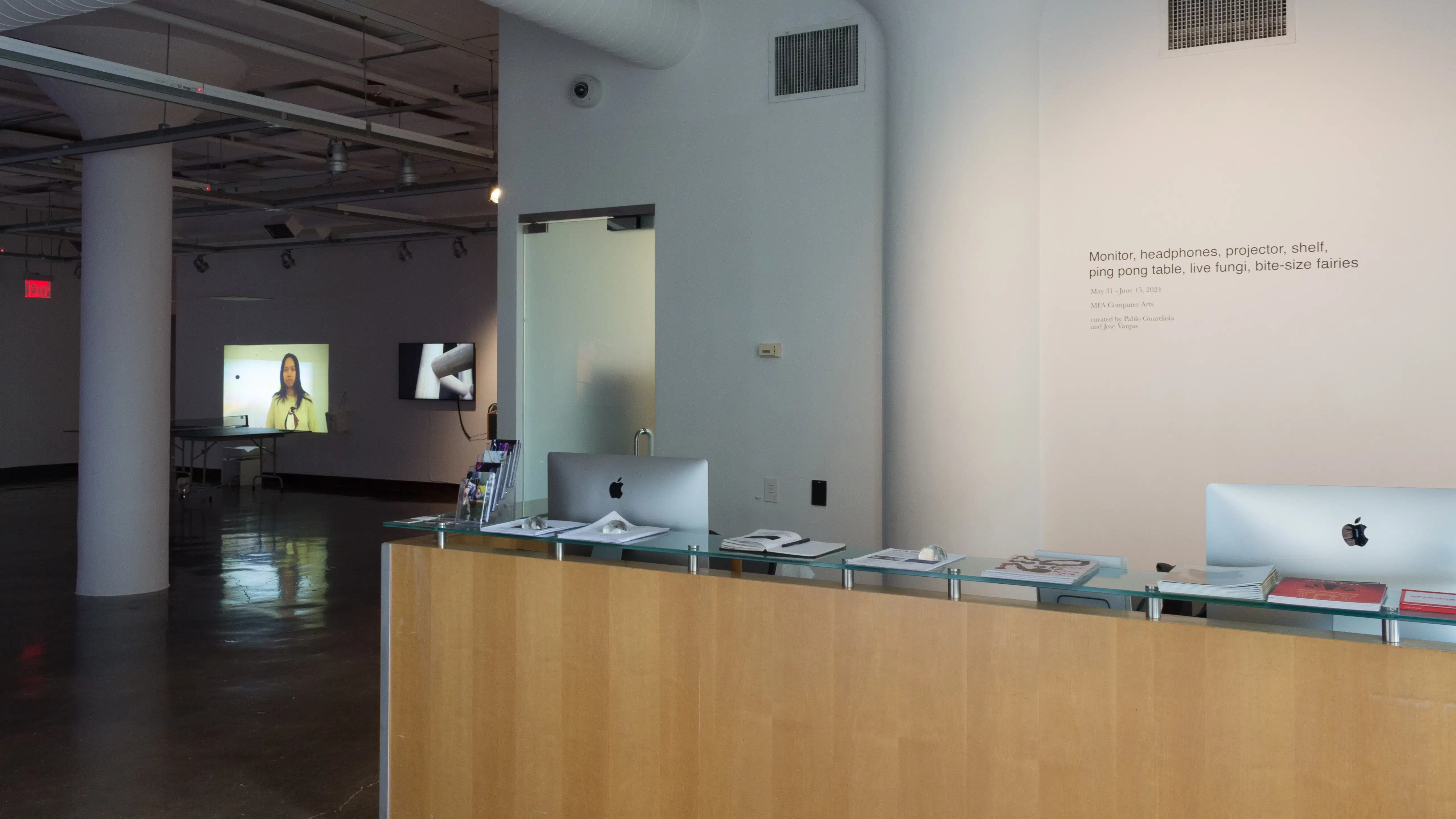Installation image showing the title wall behind a computer desk, with a large gallery in the background to the left.