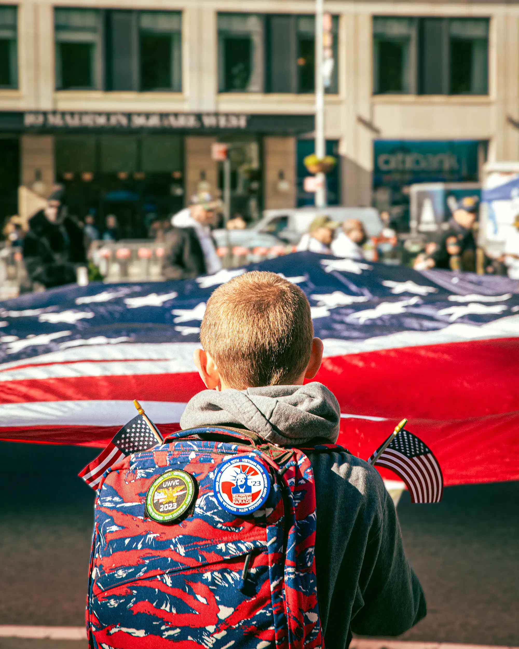 A kid faces away from the camera to look at a large american flag. He has patches on his backpack and holds two small american flags.
