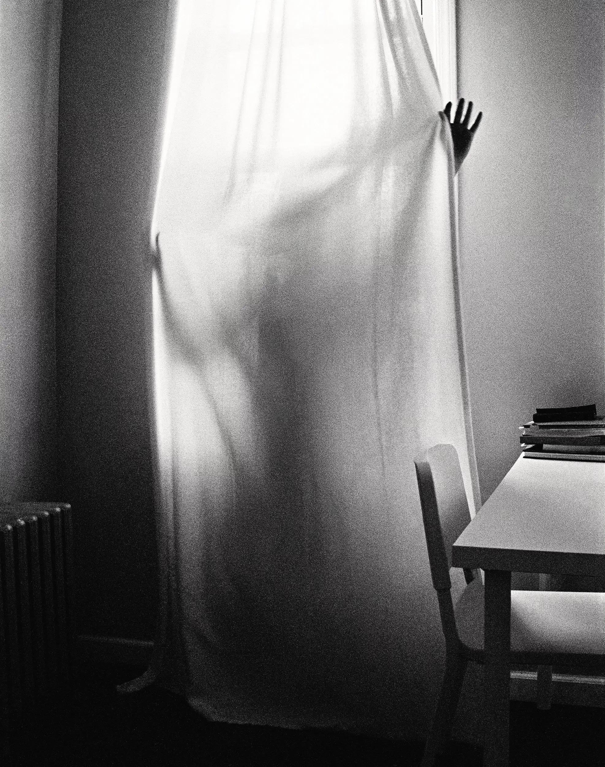 A black and white photo of someone behind a window curtain. The light coming from the window outlines the person's body.