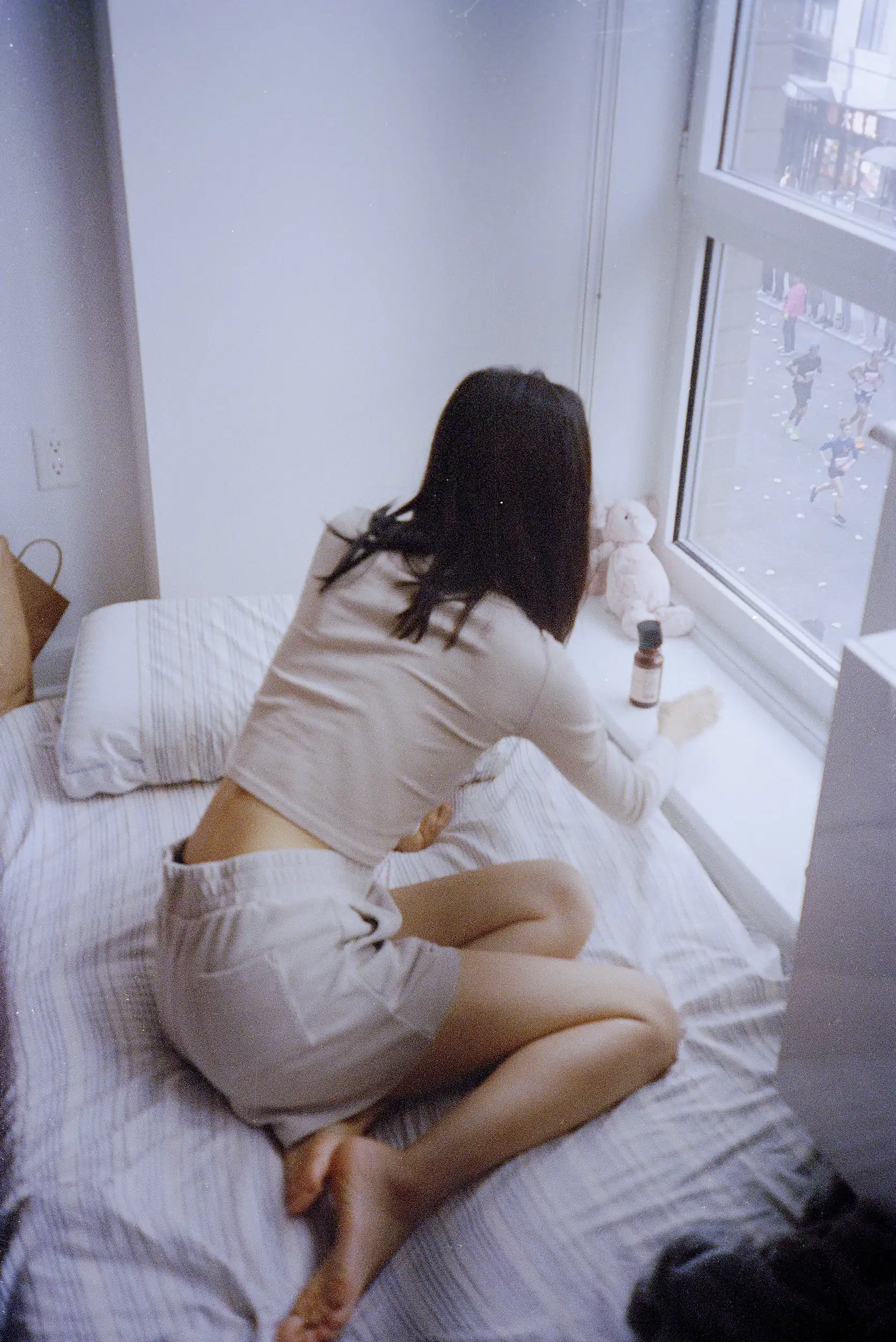 A girl sits on a bed looking out the window at a street scene below, with a soft toy and a bottle nearby.