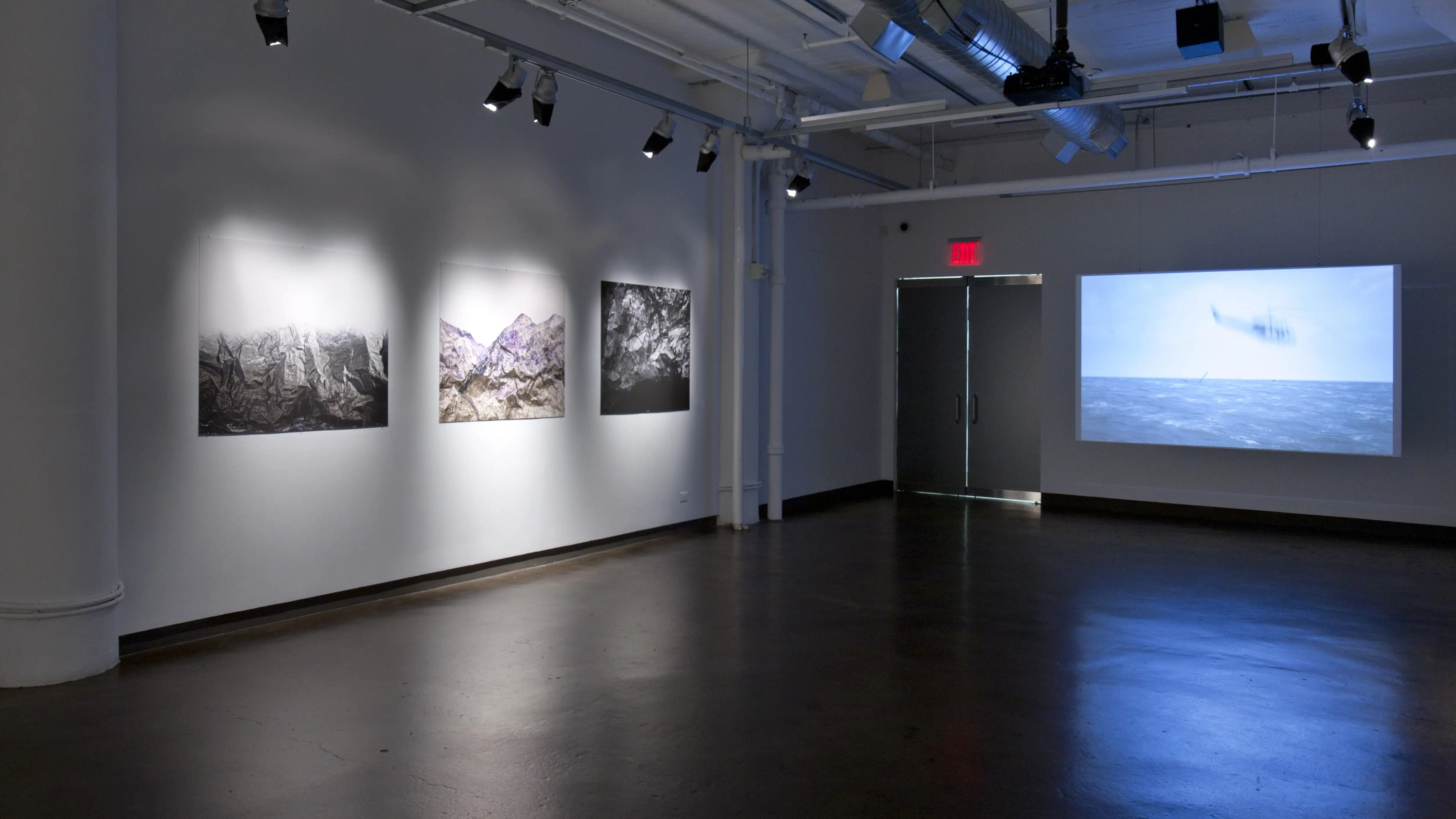 This is an art gallery with empty floor space and artwork hanging on the wall. There is a doorway in the corner with an exit sign above it.