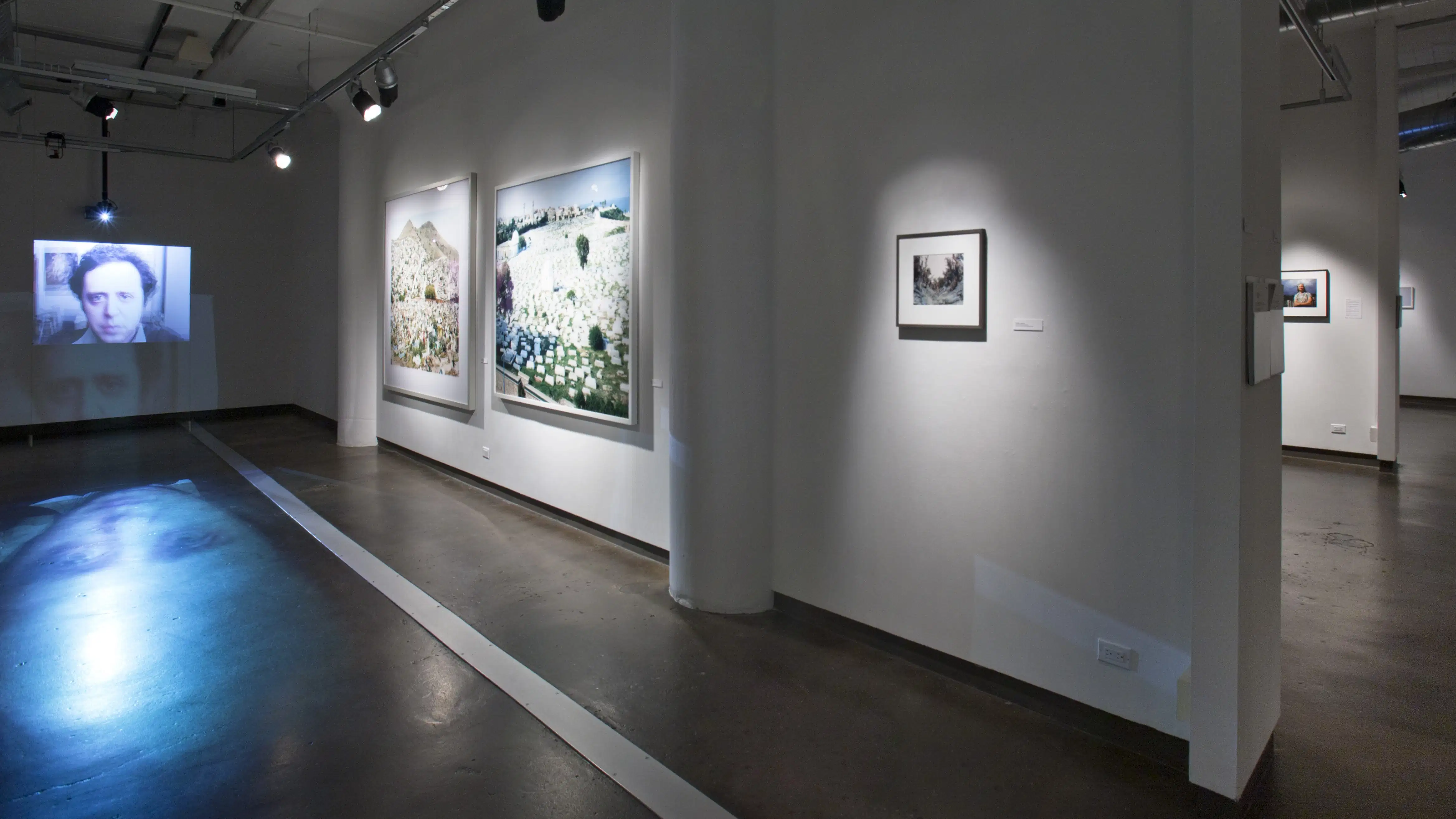 A white room with pieces of art paintings and a visual televised image of a man reflected onto the floor.