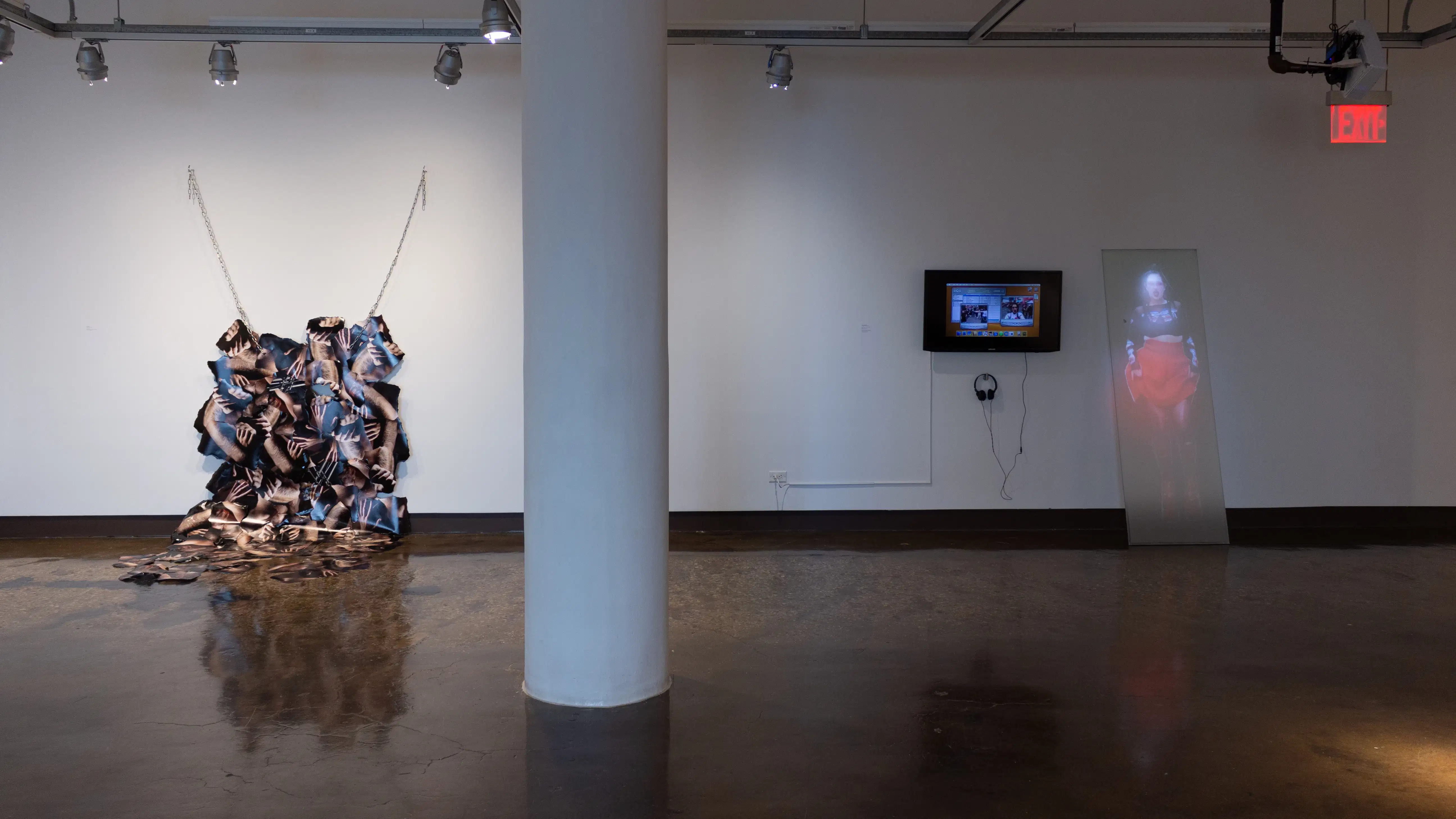 Installation image showing a column dividing the room between a hanging photo installation and a monitor and projection on a leaning glass panel.