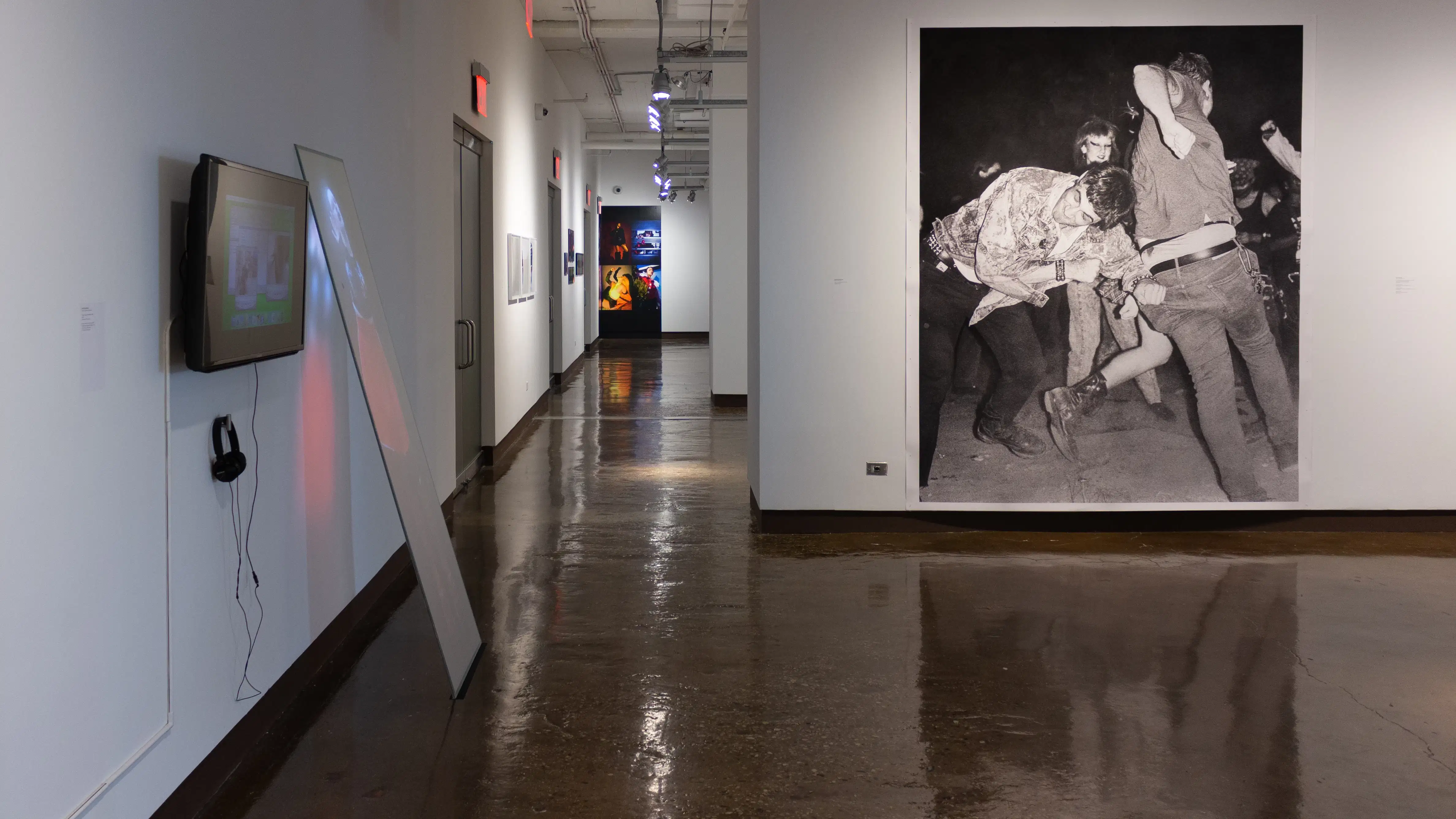 Installation image showing a monitor and projection on a leaning glass panel on the left, a view down a long hallway, and a large black and white photograph of people dancing on the right.